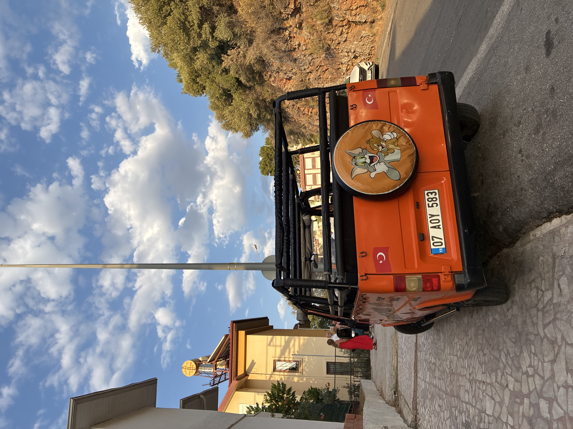 In Alanya, Turkey, an orange, open-top vehicle is parked on a paved street beside a building. The vehicle has a spare tire cover featuring Tom and Jerry cartoon characters. A Turkish flag is affixed to the rear of the vehicle. The license plate reads "07 AOY 583". Several people are standing near the building. The background shows a rocky hillside covered with vegetation and a cloudy sky.
