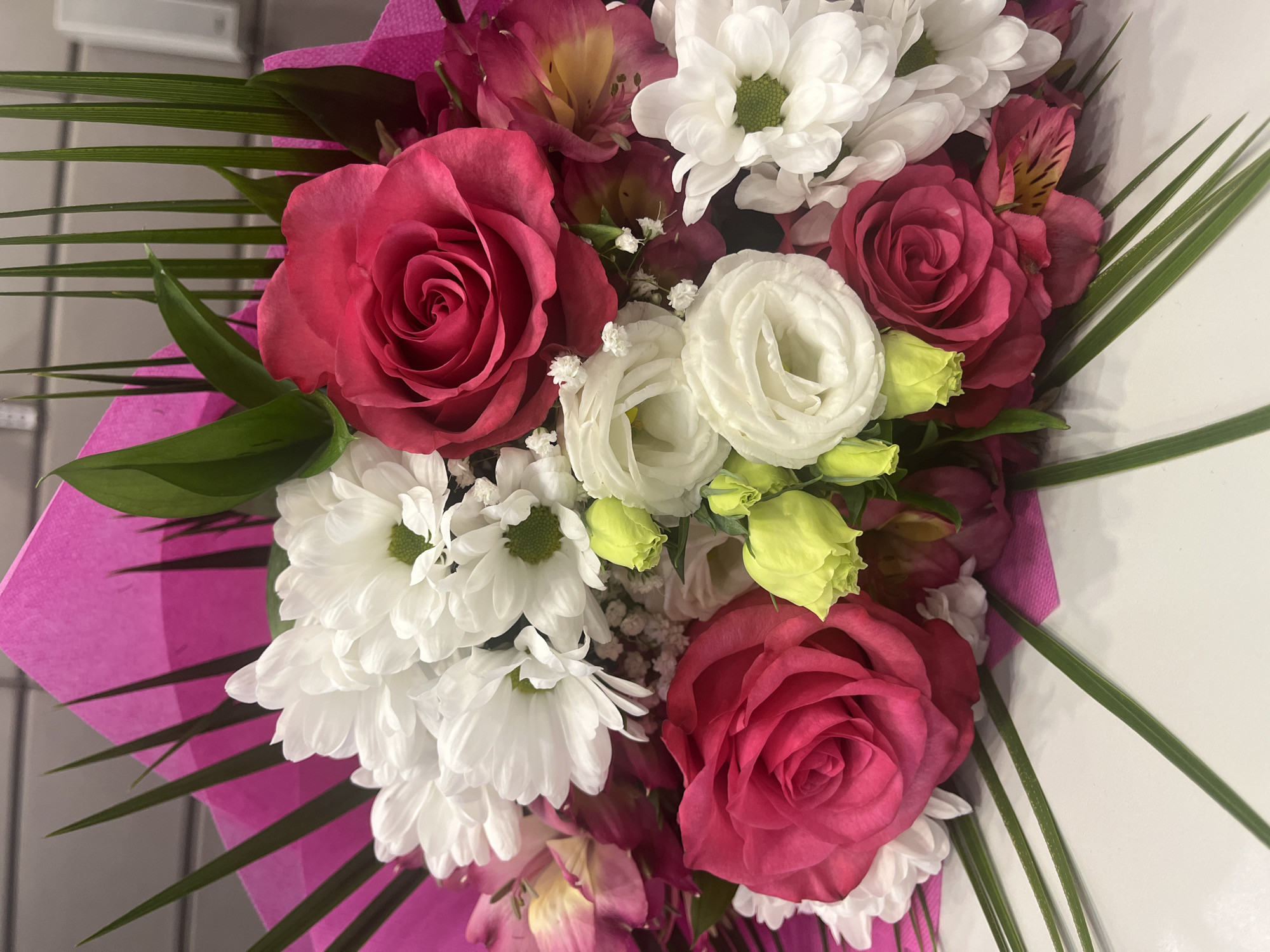 A close-up of a bouquet of flowers. The arrangement includes red roses, white chrysanthemums, white lisianthus, and pink alstroemeria, arranged with green foliage and wrapped in pink paper. The location in Plovdiv, Bulgaria is not discernible from the image alone.