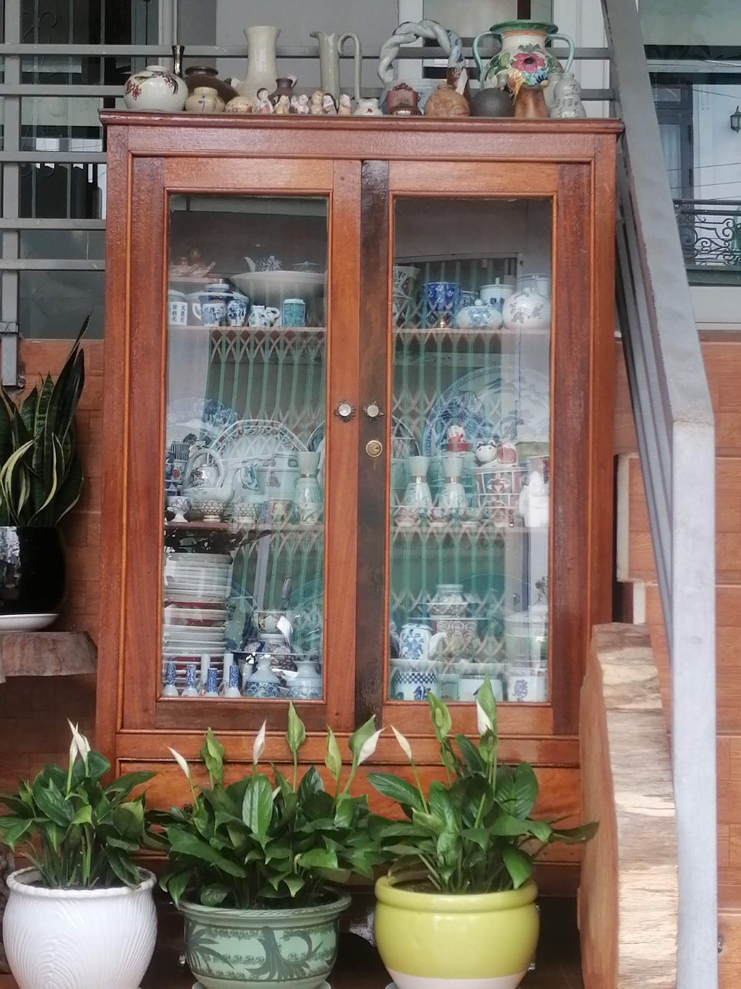 A medium shot depicts a wooden china cabinet filled with various porcelain pieces, likely located in Da Lat, Vietnam. The cabinet is situated outdoors on a tiled surface. In front of the cabinet are three potted peace lilies, the pots being white, green, and yellow. Atop the cabinet are decorative items: small figurines, vases, and pots. The cabinet's glass doors reflect the surrounding environment. To the left of the cabinet is a snake plant in a black pot. To the right is part of a staircase. The background is somewhat blurred, but appears to be an urban environment. No people are visible.