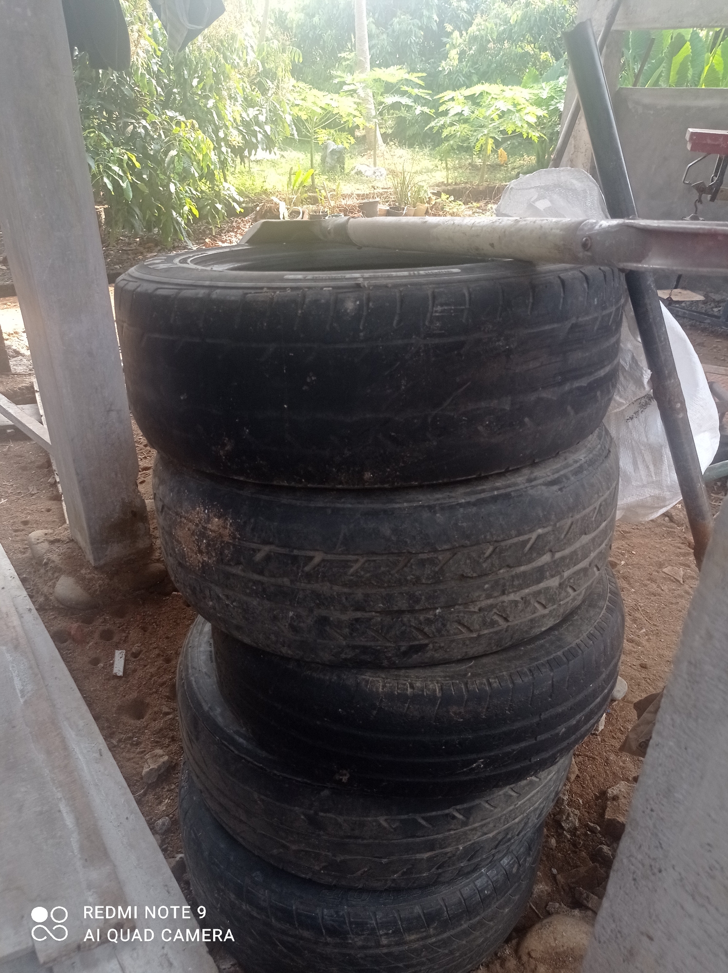 A stack of five used tires is visible outdoors, with a shovel resting on top of the highest tire. The tires are black and appear worn. Behind the tires, lush green foliage and trees are present, suggesting a natural or semi-rural environment. The ground is covered in dirt and small stones. A light-colored, textured surface, possibly concrete or a similar material, is present on the left side of the frame, and a portion of a larger, lighter structure is visible on the right. The lighting indicates it is daytime, with sunlight filtering through the trees. The scene is located in Cipawon, Indonesia, as indicated by the "REDMI NOTE 9 AI QUAD CAMERA" watermark in the bottom left corner, which is a common watermark for Xiaomi phones manufactured in or for the Indonesian market.