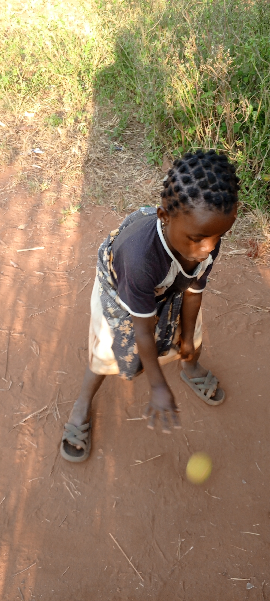 An African child in Ngalula, Democratic Republic of the Congo, stands on a red dirt path. The child wears a dark shirt with a white collar, a patterned skirt, and sandals. The child is looking down at a yellow ball, possibly a tennis ball, and appears to be in the act of bouncing the ball. The path is bordered by green vegetation on one side and dry grass on the other. Shadows are cast on the ground, suggesting sunlight.