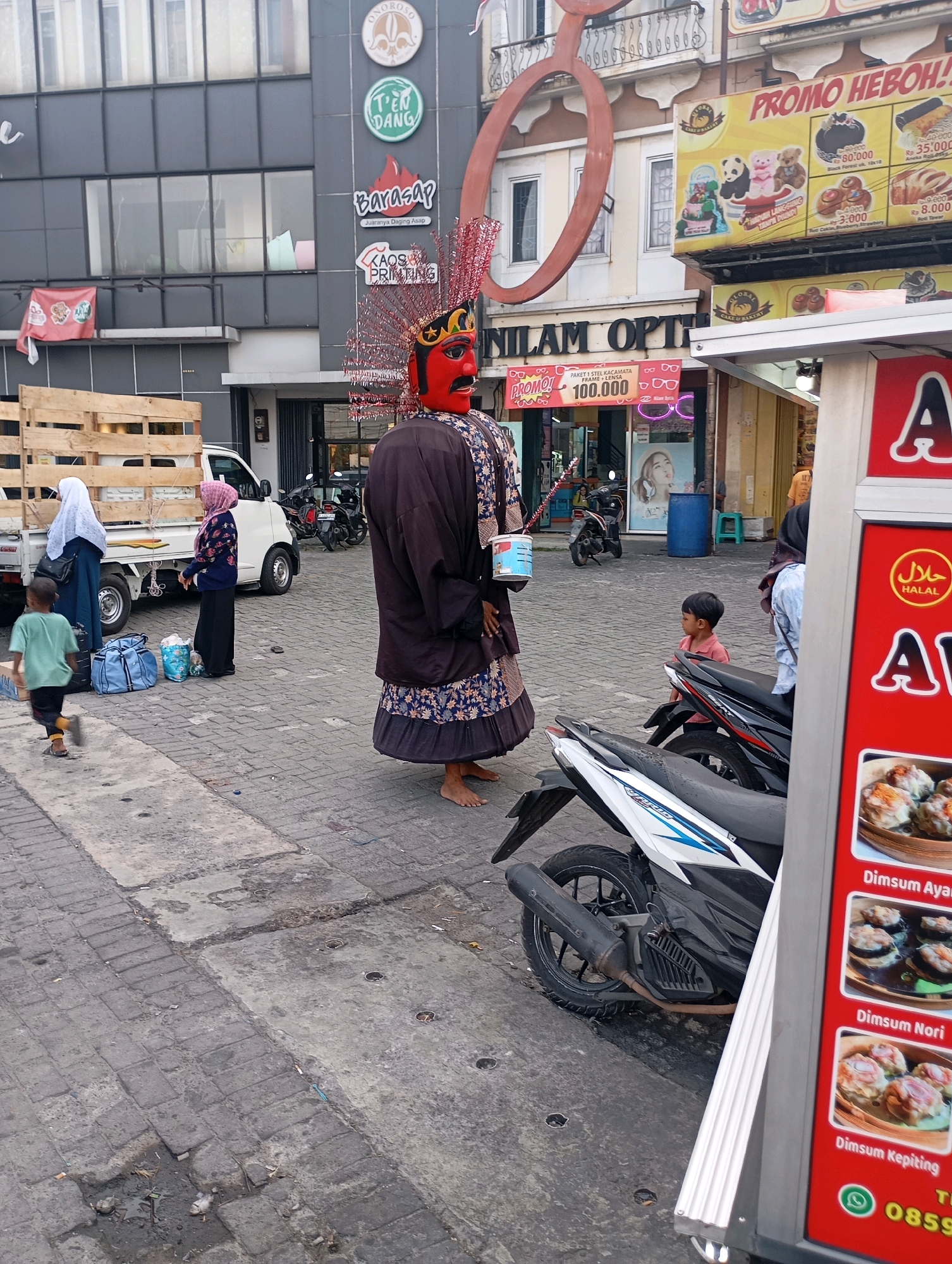 In Tangerang, Indonesia, a street scene unfolds with a variety of activities and elements. A large, traditional Indonesian 'ondel-ondel' puppet, dressed in a dark robe with a red and gold headdress, stands prominently in the foreground, holding a white bucket. To the left, a small white truck is parked, with its wooden bed loaded. Two women, one in a white hijab and blue outer garment, and another in a pink hijab and patterned jacket, stand near the truck. A young boy in a green shirt is walking away from them towards the left.
Behind the 'ondel-ondel', a building features several storefronts. One sign reads "NILAM OPTIK". Above, banners display advertisements for "PROMO HEBOH!!" and "GLOBAL CAKE & BAKERY", showcasing images of cakes and pastries. Other signs indicate businesses such as "Barasap" and "KAOS PRINTING". Several motorcycles are parked in the area.
To the right, a red food stall is visible, displaying images of dim sum with labels "Dimsum Ayam", "Dimsum Nori", and "Dimsum Kepiting". A young boy in a red shirt sits on a black motorcycle. The ground is paved with cobblestones and concrete slabs. In the background, other buildings and trees are present, contributing to the urban environment of Tangerang.