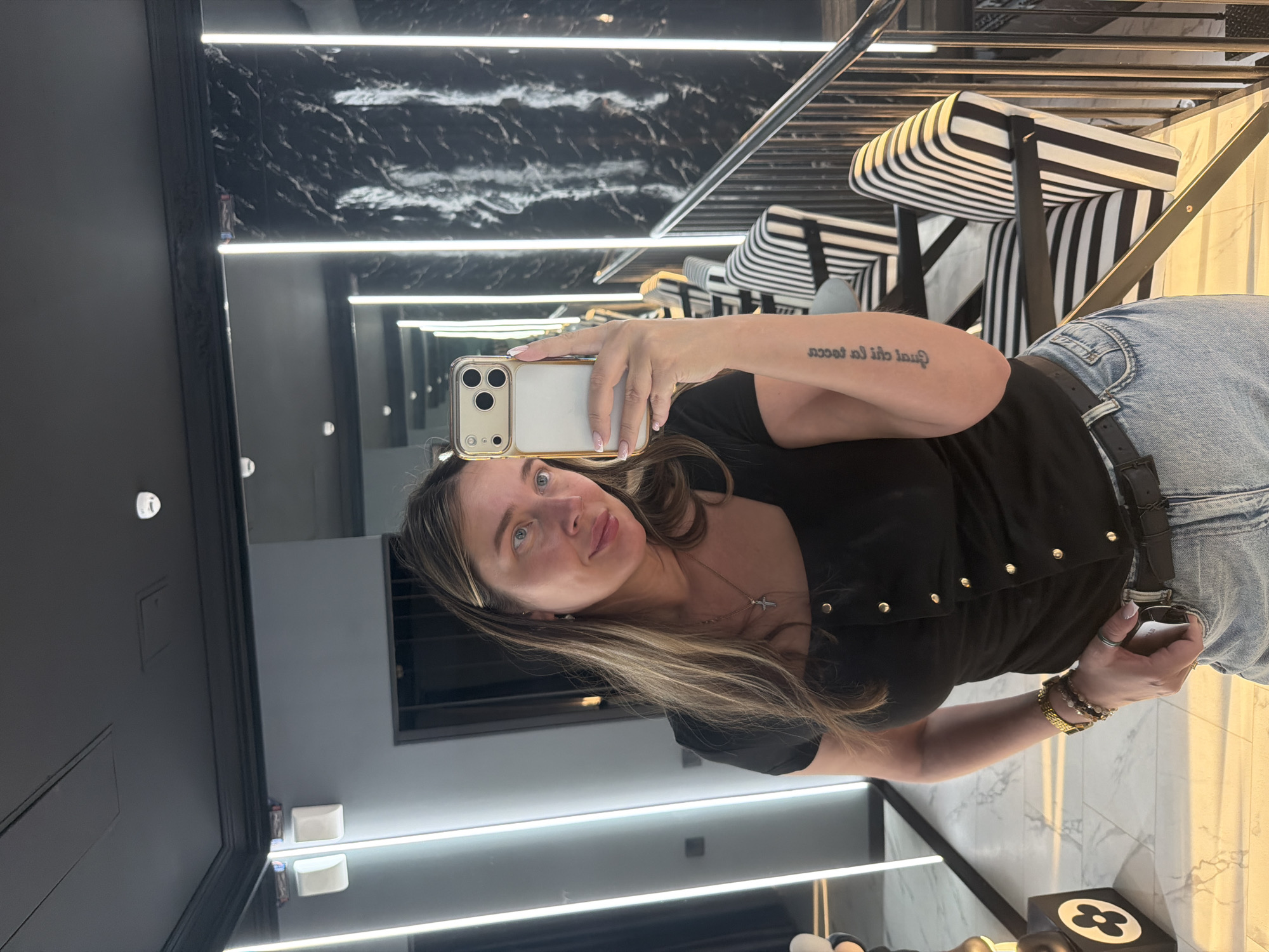 An adult female is depicted standing in a modern, upscale interior, taking a mirror selfie with a smartphone in the city of Укромновское сельское поселение, Ukraine. The woman, appearing to be in her late 20s to early 40s, has long, light brown hair with blonde highlights, parted on her left, and blue eyes. She is wearing a black short-sleeved top with a square neckline and a vertical row of gold buttons down the front. Her lower attire consists of light blue denim jeans secured by a black belt with a silver buckle.
Her left wrist is adorned with multiple beaded bracelets and a gold-toned watch. A silver cross pendant hangs from a gold chain around her neck. A ring is visible on her right hand. On her right forearm, a tattoo of script, possibly in a non-Latin alphabet, is visible. She holds a gold and white smartphone, equipped with a triple-lens camera array, in her right hand, positioned to capture her reflection in a large mirror. She exhibits a subtle smile, looking towards the phone.
The environment is characterized by dark, sleek finishes. The ceiling is black, featuring recessed lighting and rectangular ventilation grilles. Walls are a mix of dark grey panels and black panels with a marble-like pattern, illuminated by prominent vertical LED light strips. The floor is light-colored, possibly polished stone or tile, displaying a subtle geometric pattern. In the background on the right, several chairs with distinctive black and white vertical stripes on their upholstery and black frames are visible. A metal railing separates this seating area, suggesting a change in elevation or a mezzanine. On the far left, a dark square pedestal with a white circular logo resembling a stylized four-petal flower is present. Two white, square-shaped fixtures are mounted on the upper left wall. The overall lighting is bright and evenly distributed, emanating primarily from the vertical LED strips.