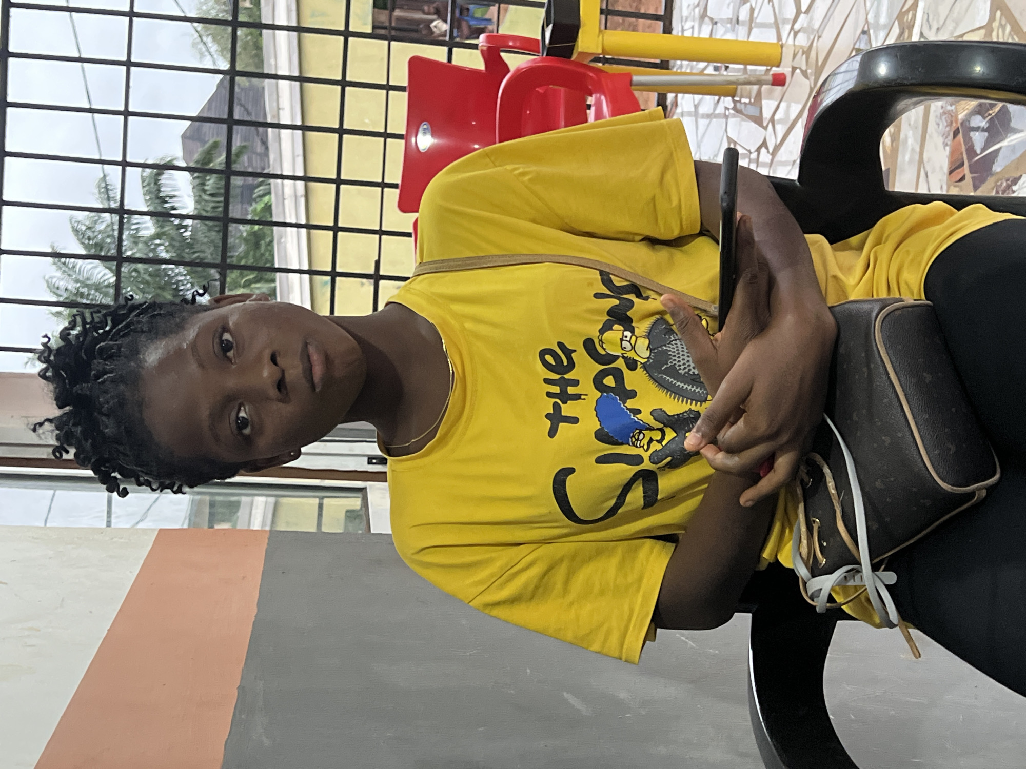A young Black female sits in a black plastic chair facing the camera. She has dark skin, black braided hair styled in twists, and is wearing a yellow t-shirt with a graphic of Homer Simpson from The Simpsons. She has a dark brown purse with a tan strap slung across her chest. Her hands are clasped in front of her, and she is holding a black smartphone. The background shows a wall with horizontal bands of orange and gray, a window with a black metal grate, and outdoor elements including palm trees and a red plastic chair. The scene appears to be in Nkpor, Nigeria, likely in a residential or commercial area, given the mixture of indoor and outdoor elements and visible structures.