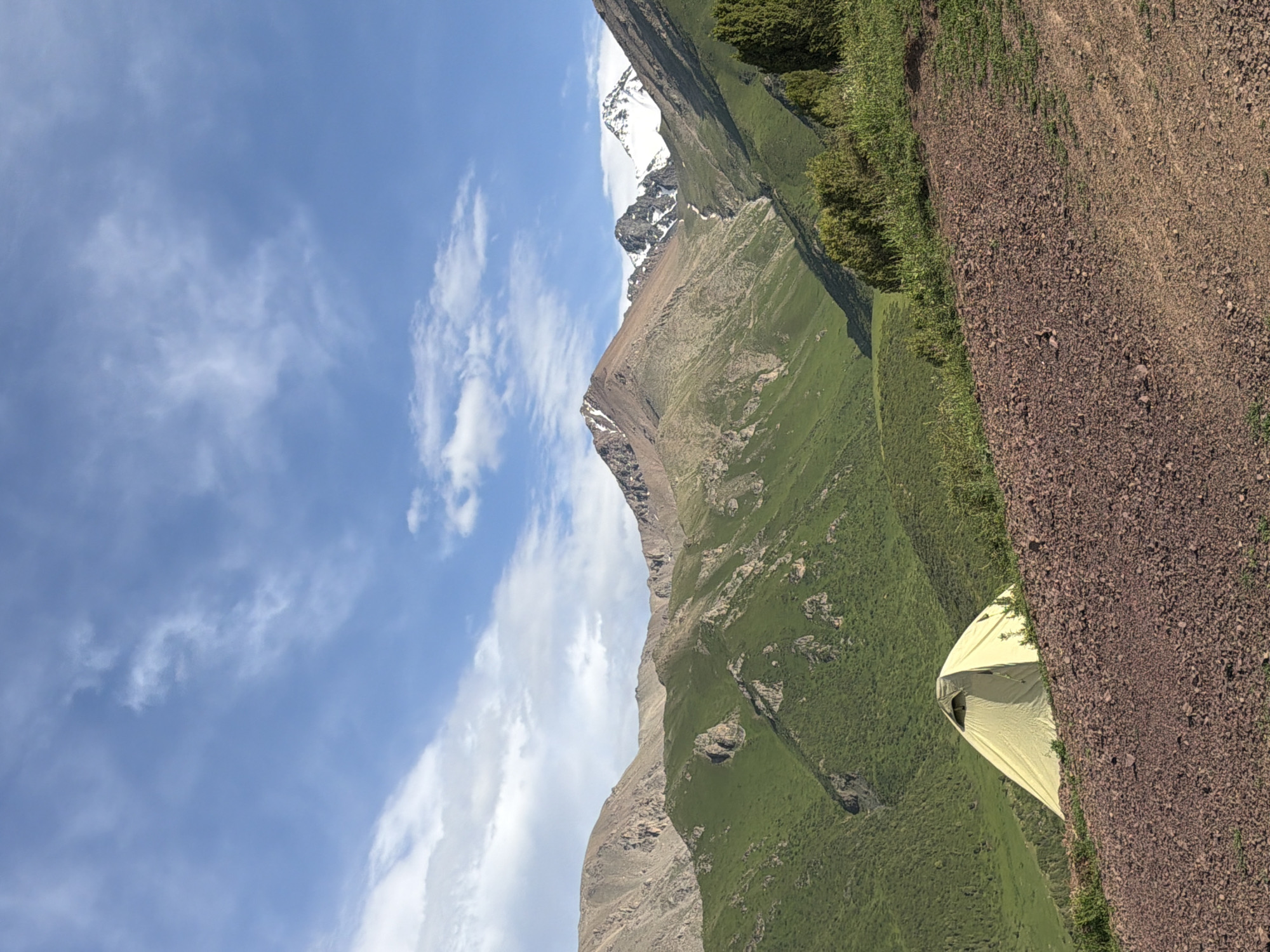 A light yellow tent is pitched on a grassy slope in front of a vast mountain range under a partly cloudy blue sky. The mountains in the background are covered in green vegetation and rocky outcrops, with some peaks showing snow. The foreground is a dirt path with small rocks and sparse green grass. The image appears to be taken in a natural, outdoor setting, possibly in the region of Kashka Suu, Kyrgyzstan, given the terrain and vegetation. There are no people visible in the image.
