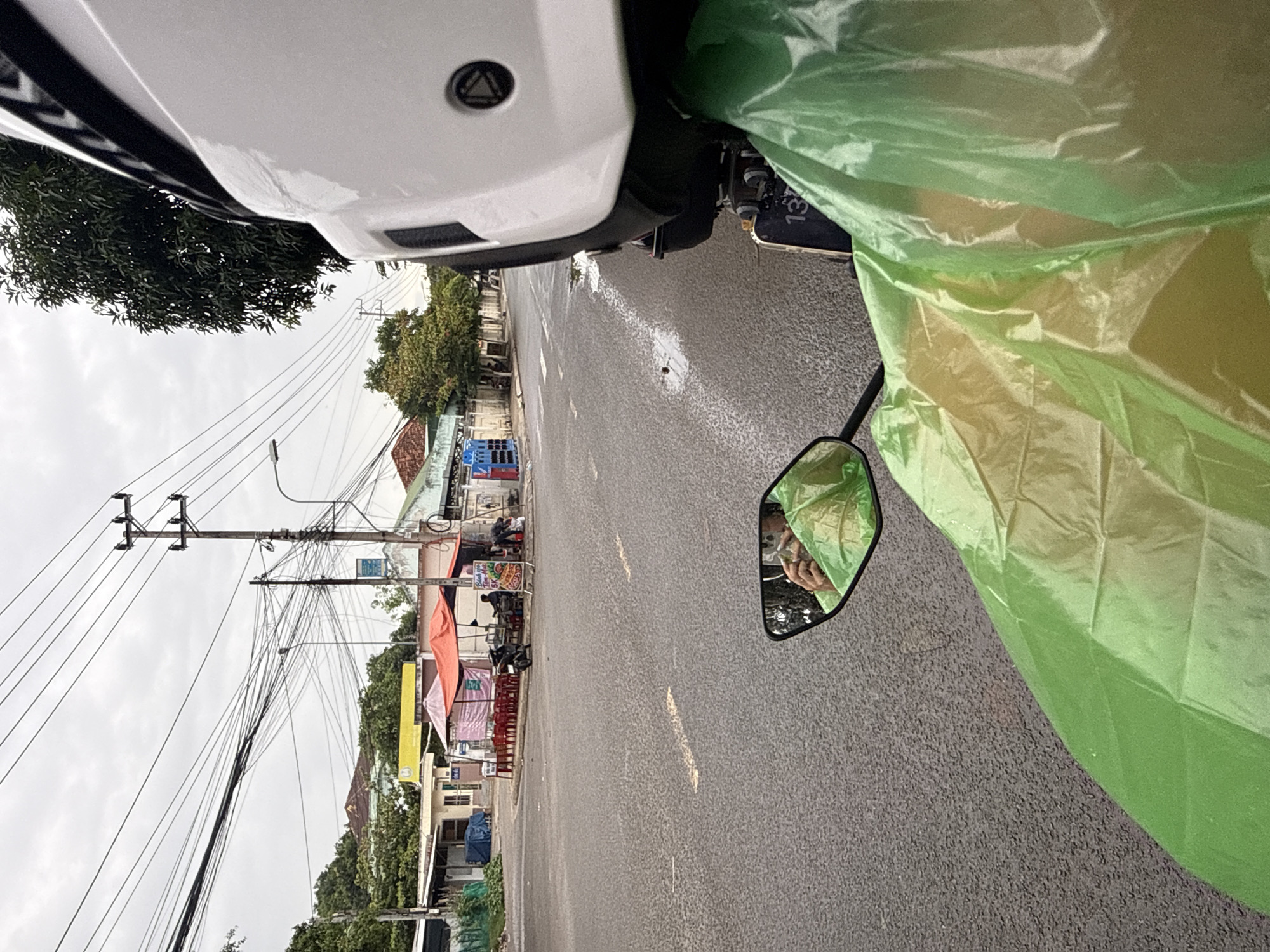 This is a first-person perspective photograph taken from a motorcycle in Nha Trang, Vietnam. The road surface is wet, indicating recent rain, and there are puddles visible. In the foreground, a bright green poncho covers the rider and part of the motorcycle. A side mirror reflects the rider's hands, also covered by the poncho. The background shows a street scene with several small shops or food stalls, some with orange and pink awnings, and red plastic chairs set up outside. Electrical wires are strung across the sky, supported by utility poles. A person is seen working at one of the stalls. The overall atmosphere is overcast, suggesting a cloudy day.