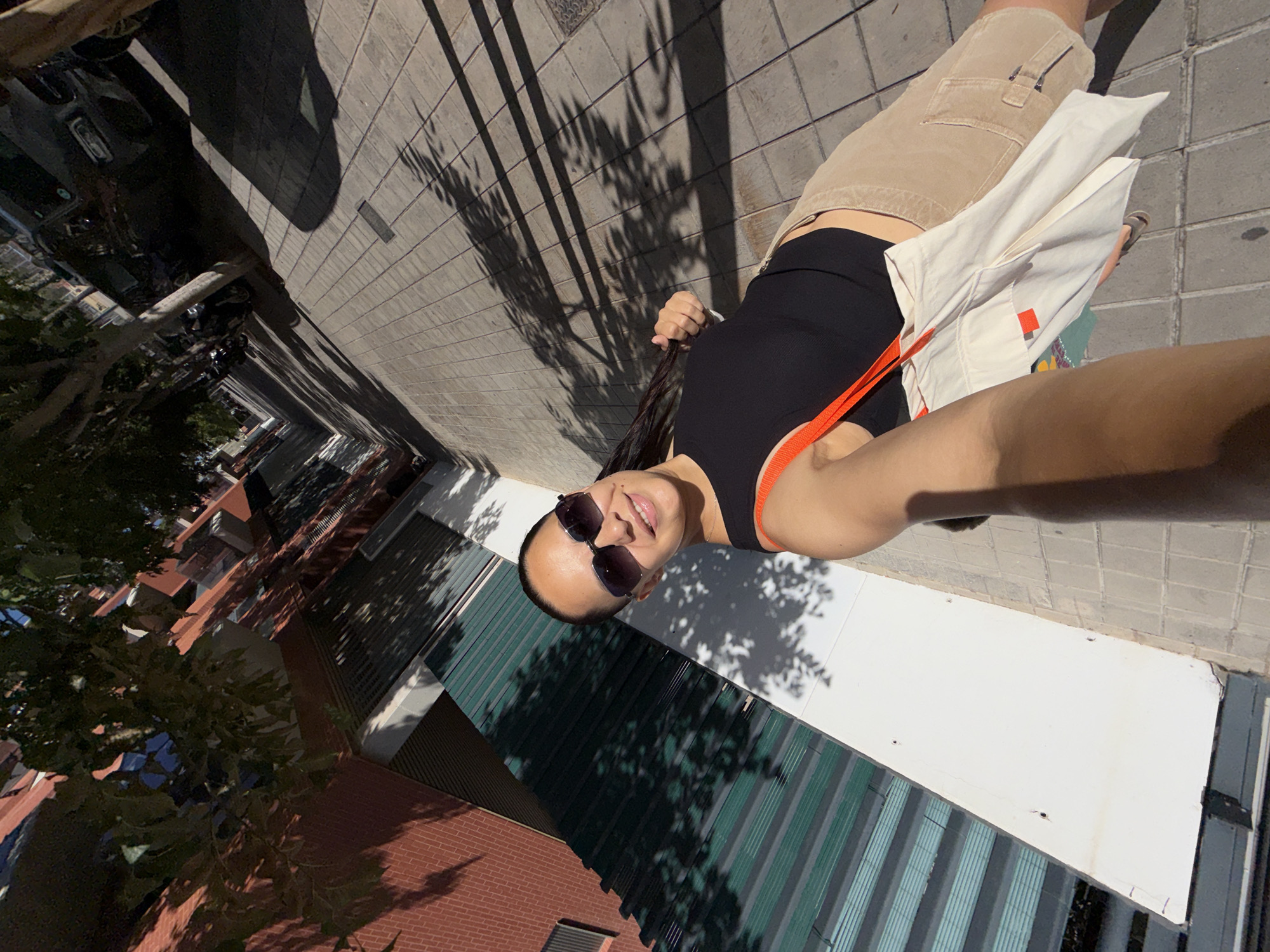 In a high-angle, full-body selfie taken on a sidewalk in an urban environment, a woman is captured from the chest up, holding the camera with her left arm, which is extended forward. She is wearing dark sunglasses and a black tank top with orange accents. Her right hand is in her hair, which is dark and long, with some strands falling forward. She is also wearing khaki shorts and carrying a white tote bag with orange straps, which is slung over her shoulder. The woman is smiling at the camera.
The background reveals a perspective view of a city street with a brick building on the left, featuring green blinds and windows. Trees with dappled sunlight cast shadows on the sidewalk, which is paved with rectangular tiles. In the distance, cars and motorcycles are parked along the street. The scene suggests a bright, sunny day in an urban setting, possibly a residential or commercial area.