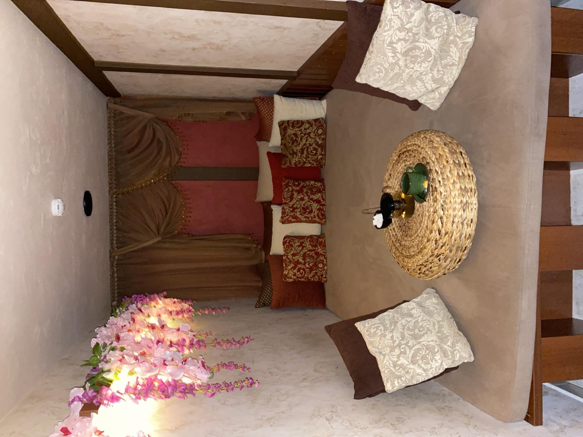 The image depicts a cozy, dimly lit room with a Middle Eastern or Moroccan ambiance. The primary subject is a low seating area furnished with a large, plush couch and numerous decorative pillows. These pillows vary in color and pattern, with some being a deep red adorned with intricate gold designs, and others featuring a neutral cream color with an embossed floral pattern. Adjacent to the couch is a round, woven basket-like coffee table. On this table, there's a small green teacup and saucer, along with what appears to be a tea set.
The room has a rustic ceiling with exposed wooden beams. To the left, a wall is painted in a light, textured finish, and a white circular object, possibly a smoke detector, and a black circular object, are mounted on it. Further to the left, a cascade of artificial pink and purple flowers is arranged, adding a touch of color and vibrancy to the scene.
The lighting in the room is soft and warm, creating a relaxed and inviting atmosphere. The overall impression is one of comfort and tranquility, suggesting a space designed for relaxation or intimate gatherings. The style and decor imply a possible spa, lounge, or a private relaxation room. No people are visible in the image. The time of day is indeterminate due to the interior setting and controlled lighting. There is no visible text in the image.