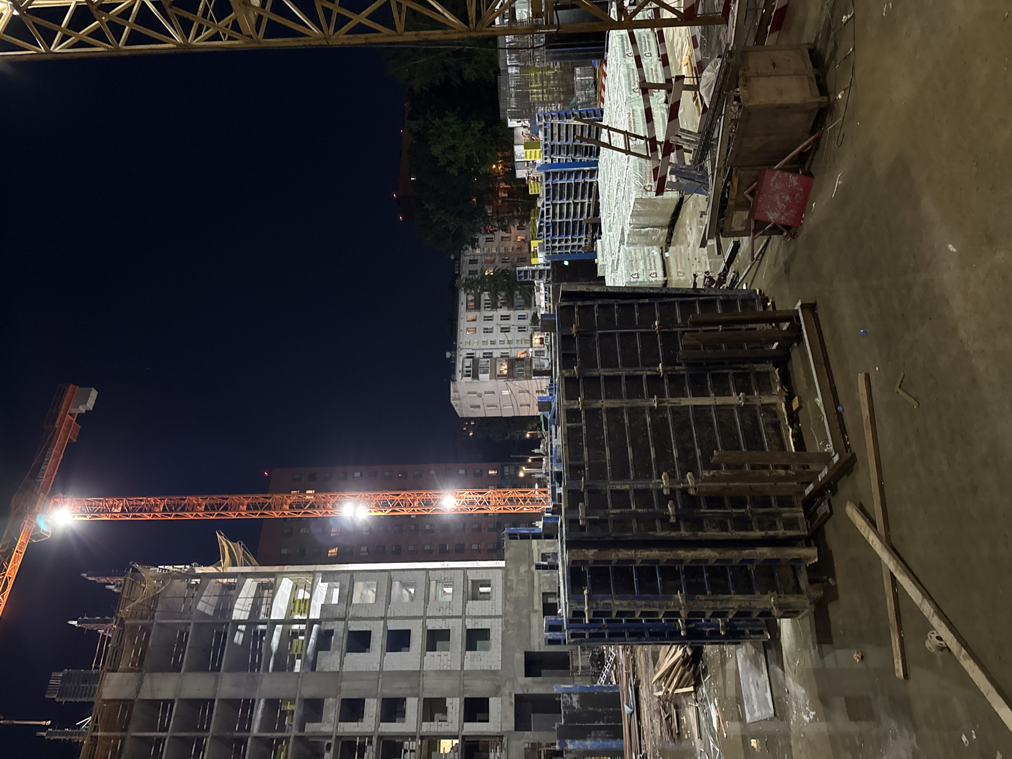 This is an eye-level, medium shot of a construction site at night in Moscow, Russia. The scene is illuminated by the bright lights of a construction crane and lights on the unfinished building, casting long shadows. In the foreground, there are large, dark grey construction forms, possibly for concrete pouring, with blue metal accents. To the left, a partially constructed multi-story building, made of concrete and cinder blocks, rises into the night sky. The building is skeletal, with open window frames and exposed rebar. An orange construction crane with its boom extended dominates the upper left portion of the image, with lights illuminating the structure. In the midground, to the right of the crane, a red brick residential building is visible, with lights on in some windows. Further back, several other residential buildings can be seen, their facades illuminated. In the midground and right, there are stacks of building materials, including pallets of white-wrapped items that appear to be insulation or building boards, and a metal ladder leaning against them. The ground is wet and reflective, suggesting recent rain. Wooden planks and other construction debris are scattered across the wet concrete surface. The overall atmosphere is one of ongoing construction under artificial light, with the dark night sky providing a backdrop.