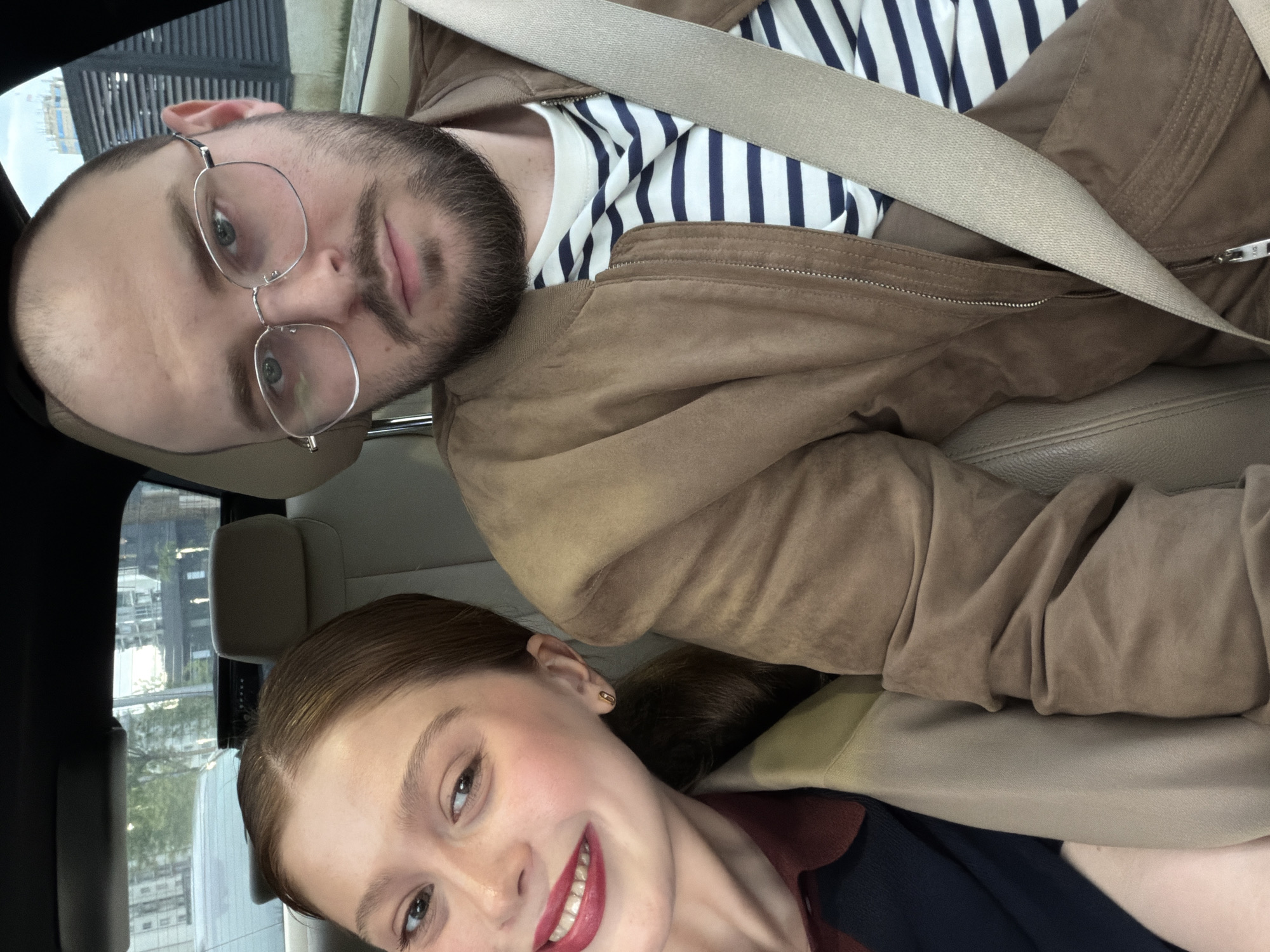 The image is a selfie taken inside a vehicle, likely a car, in what appears to be Tbilisi, Georgia. A man and a woman are visible. The man, positioned at the top of the frame, has a beard, wears glasses, and is wearing a striped shirt under a brown jacket with a seatbelt across his chest. The woman, positioned lower in the frame, has red hair, is smiling, and wears a dark top underneath a beige coat. The car's interior features beige leather seats and dark trim. Outside the window, buildings are visible, suggesting an urban environment.