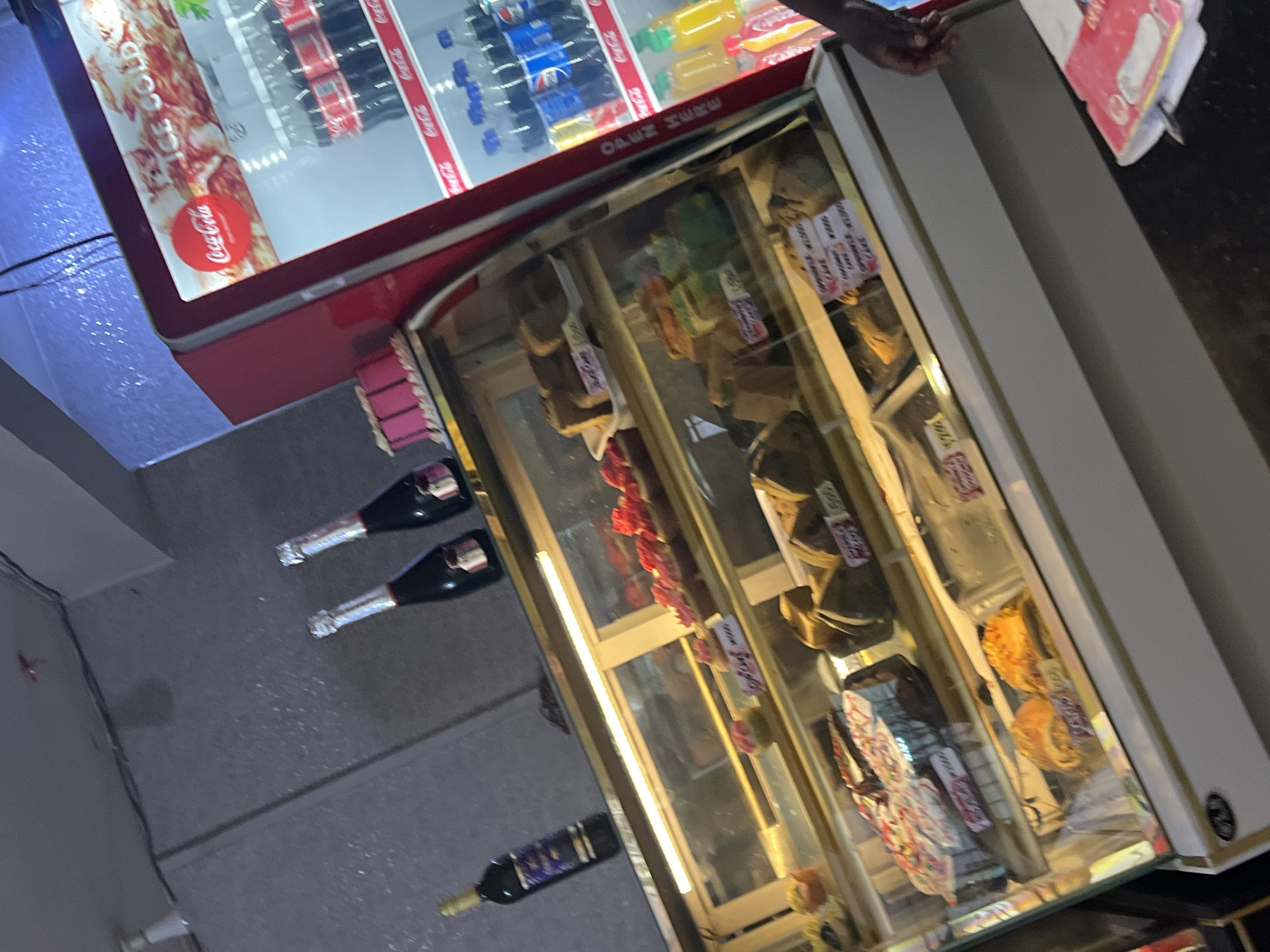 The image shows a refrigerated display case, likely in a shop or store in Jos, Nigeria. The top section of the case contains bottles of Coca-Cola and other beverages. Below, a glass-fronted display cabinet contains pastries and other food items. Three bottles of sparkling wine lie on the floor next to the display case.
