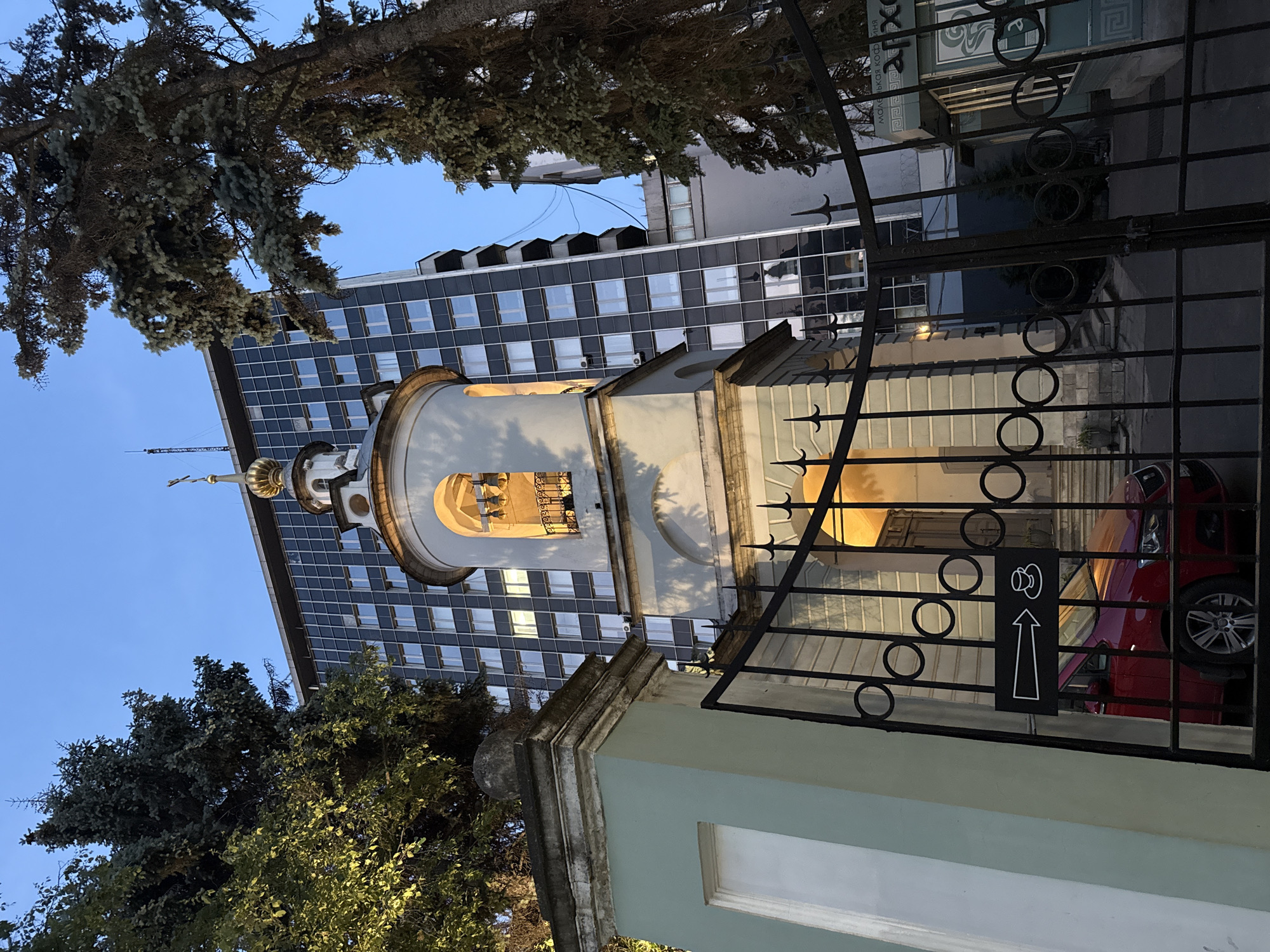 The image shows a bell tower with a golden dome and cross, in front of a modern, dark-paneled building. Trees are visible on either side, framing the scene. A black metal fence with circular designs is in the foreground, with a red car parked behind it. A sign on the fence has an arrow pointing to the right and an icon of a coffee cup. The sky is blue and clear. This is the bell tower of the Cathedral of the Archangel Michael in the Kremlin, Moscow, Russia.