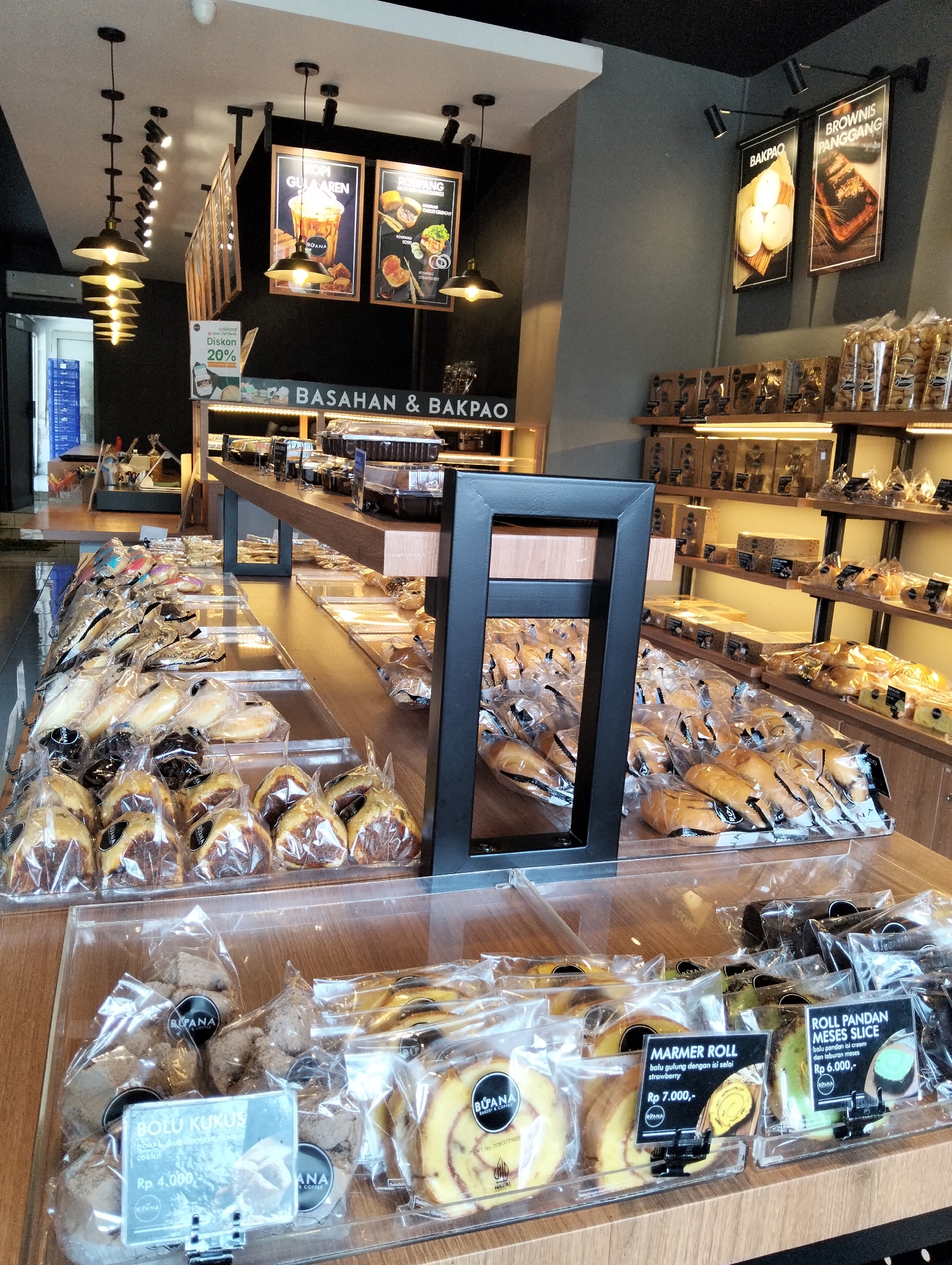 The image depicts the interior of a bakery, identified by signs as "BASAHAN & BAKPAO". The establishment appears to be located in Magelang, Indonesia, as indicated by the presence of Indonesian language text on product labels and signage. The bakery features a modern, clean aesthetic with warm lighting and wooden accents.
In the foreground, a display counter showcases a variety of baked goods, predominantly bread and cakes, neatly packaged in clear plastic. Some labels provide product names and prices in Indonesian Rupiah, such as "BOLU KUKUS coklat Rp 4.000,-", "MARMER ROLLstrawberry Rp 7.000,-", and "ROLL PANDAN MESES SLICE Rp 6.000,-". The packaging and presentation suggest a focus on hygiene and visual appeal.
Behind the display counter, more shelves are visible, stocked with additional baked items and boxed confections. Hanging from the ceiling are multiple industrial-style pendant lights, contributing to the ambient lighting. On the walls, framed posters advertise various bakery products, including "KOPI GULA AREN", "ROMPANG", "BAKPAO", and "BROWNIS PANGGANG". One poster also advertises a "Diskon 20%".
The overall environment is well-lit and organized, suggesting a commercial establishment focused on retail sales of baked goods. No individuals are present in the immediate view, allowing the focus to remain on the products and the bakery's interior design. The scene is captured from a slightly elevated perspective, looking across the display area.