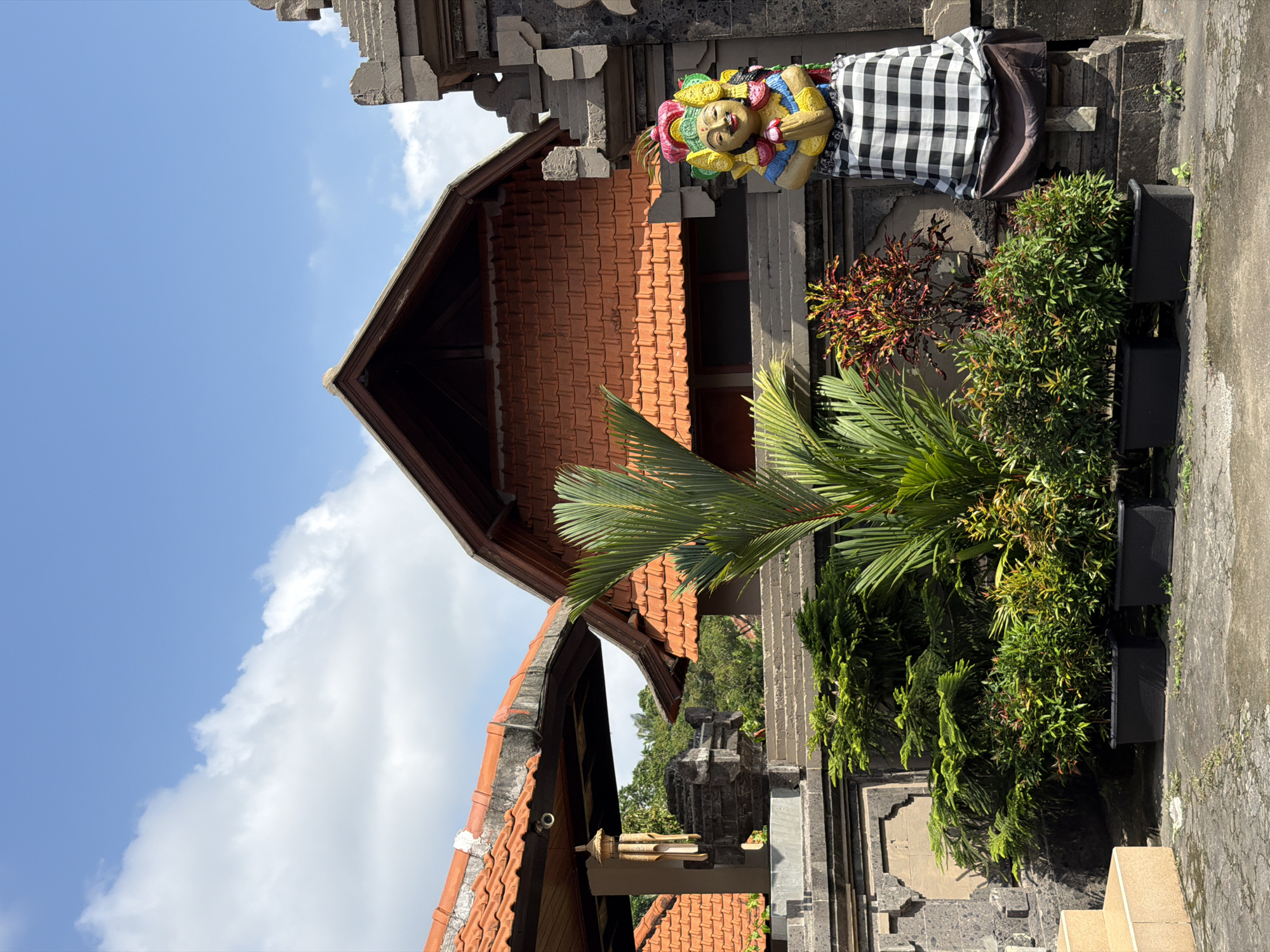This is a medium shot of a courtyard in Ubud, Indonesia. The scene is dominated by traditional Balinese architecture with red tile roofs and stone walls. In the foreground, several potted plants are arranged, including a tall palm tree and dense green bushes. To the right, a colorful statue of a figure in prayer is dressed in a black and white checkered sarong. Behind the plants and statue, a multi-level building with a reddish-brown tiled roof is visible. The sky above is clear blue with a few white clouds. The ground is paved with concrete. The overall impression is one of a serene and culturally rich outdoor space.