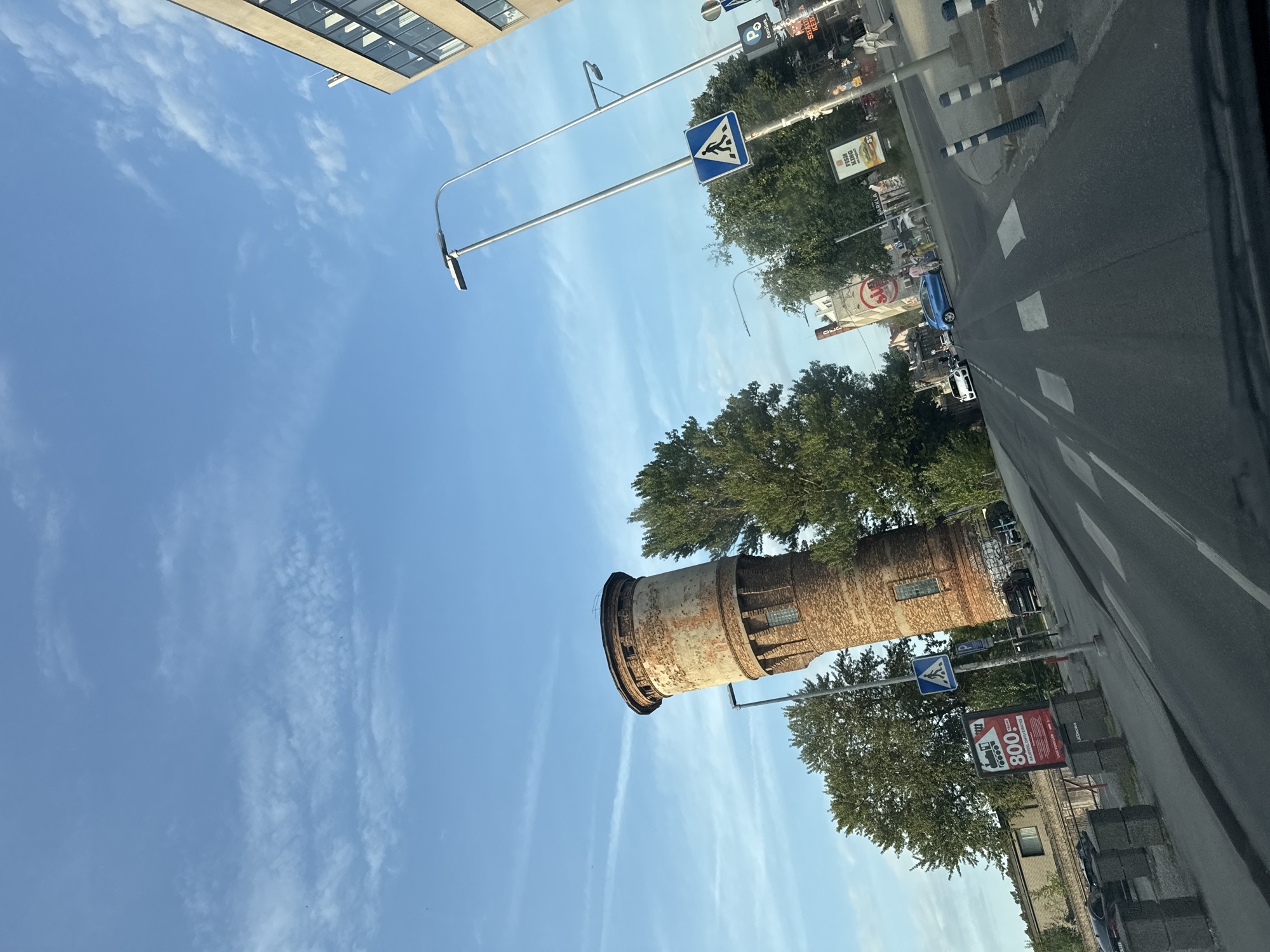 The image captures a street scene in Tallinn, Estonia, likely near the Kalamaja district, characterized by its mix of industrial heritage and modern urban elements. Dominating the left side of the frame is an old, brick water tower, its weathered facade showing signs of age. The tower stands tall against a bright blue sky with scattered white clouds.
In the foreground, a paved road with white pedestrian crossing markings leads into the distance. A blue car is visible turning on the road, and several other vehicles are present further down. On the right side of the road, a modern building with large windows and a light-colored facade stands. Streetlights and various traffic signs, including a pedestrian crossing sign, are installed along the street.
Along the left side of the road, a sidewalk area is visible with some grey concrete blocks and a large red advertisement board featuring a train illustration and the number "800". Behind this, there's a low-rise building. Trees with lush green foliage are interspersed throughout the scene, providing a natural contrast to the urban structures. The overall impression is a dynamic street view in a European city, blending historical architecture with contemporary life.