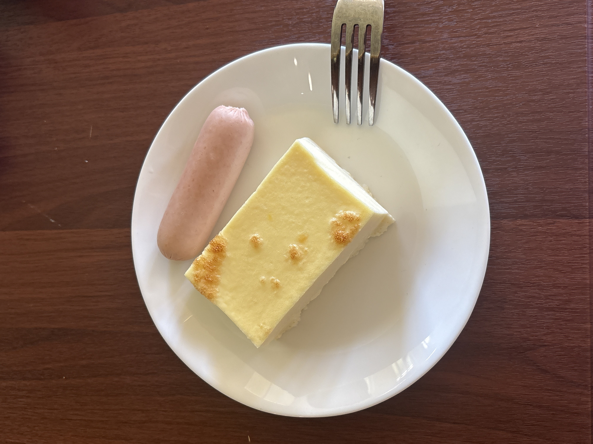 A white plate rests on a dark wooden table. On the plate are a slice of rectangular cheesecake with a golden-brown top and a pink sausage. A silver fork with four tines is positioned to the right of the plate. The scene is lit from above.