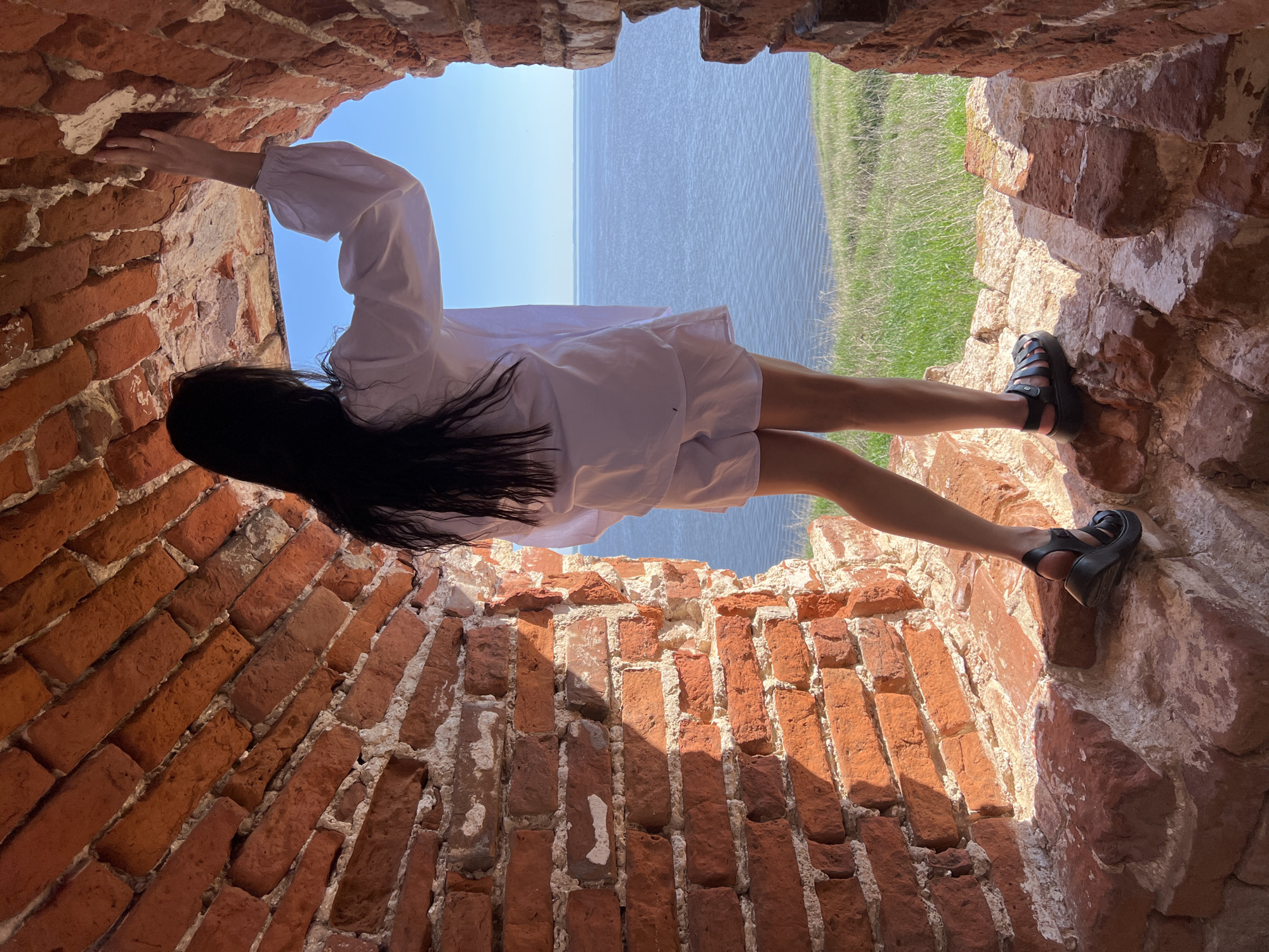 A person is seen within a brick archway, potentially a window or opening in a wall. The person is wearing a white shirt or dress and dark sandals. The person is holding onto the brick structure with one hand. The background visible through the archway includes water and grass. The location is indicated as Шуран, Russia.