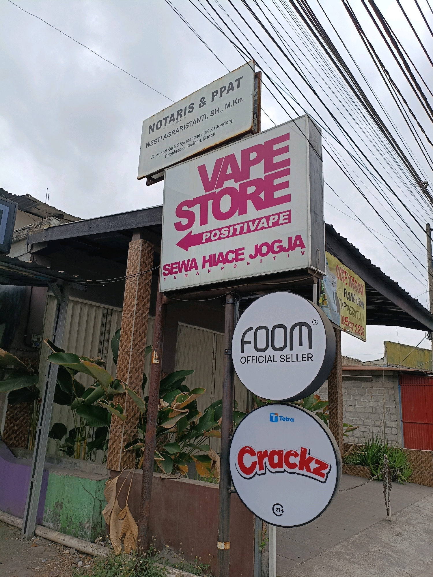The image shows a street scene in Kasihan, Bantul, Indonesia, under an overcast sky. A multi-tiered signpost is the central subject. The topmost sign, a white rectangle, reads "NOTARIS & PPAT WESTI AGRARISTANTI, SH., M.Kn." with an address below: "JL. Bantul Km 5.5 Nyemengan / DK X Glondong Tirtonirmolo, Kasihan, Bantul." Below this, a larger pink and white sign proclaims "VAPE STORE" with the tagline "POSITIVAPE SEWA HIACE JOGJA." Two circular signs hang from the post at a lower level. The upper circular sign is white with black lettering, displaying "foom OFFICIAL SELLER." The lower circular sign is also white, featuring the red text "Crackz" with a small blue Tetra logo above it, and a "21+" symbol at the bottom. A brown pole supports the signs, which are positioned in front of a building with a dark roof overhang. To the left of the signpost, lush green plants are visible. The ground in the foreground is a paved path and some rougher terrain with scattered weeds. Electrical wires are prominently strung across the top of the image. To the right, glimpses of a brick wall and a red structure are visible.