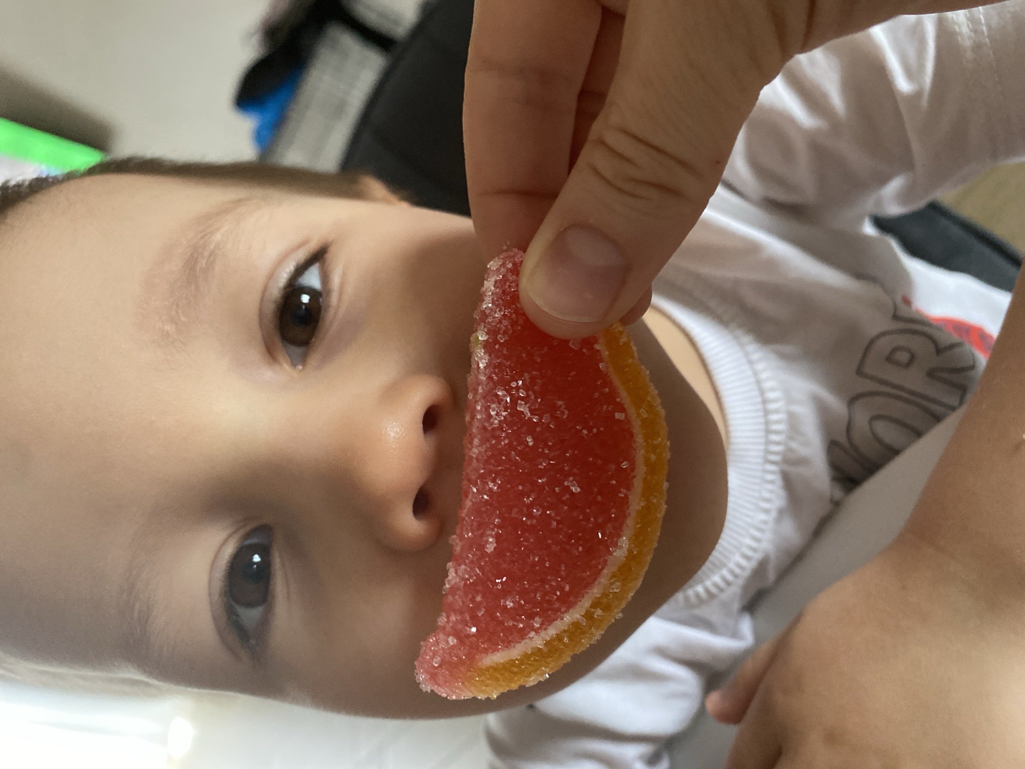 A young child is lying down wearing a white t-shirt. A hand is holding a red and yellow wedge-shaped candy near the child's face. The candy appears to be covered in sugar. The location is not provided in the image, and thus I cannot specify that the image was taken in Chaikovsky, Russia.