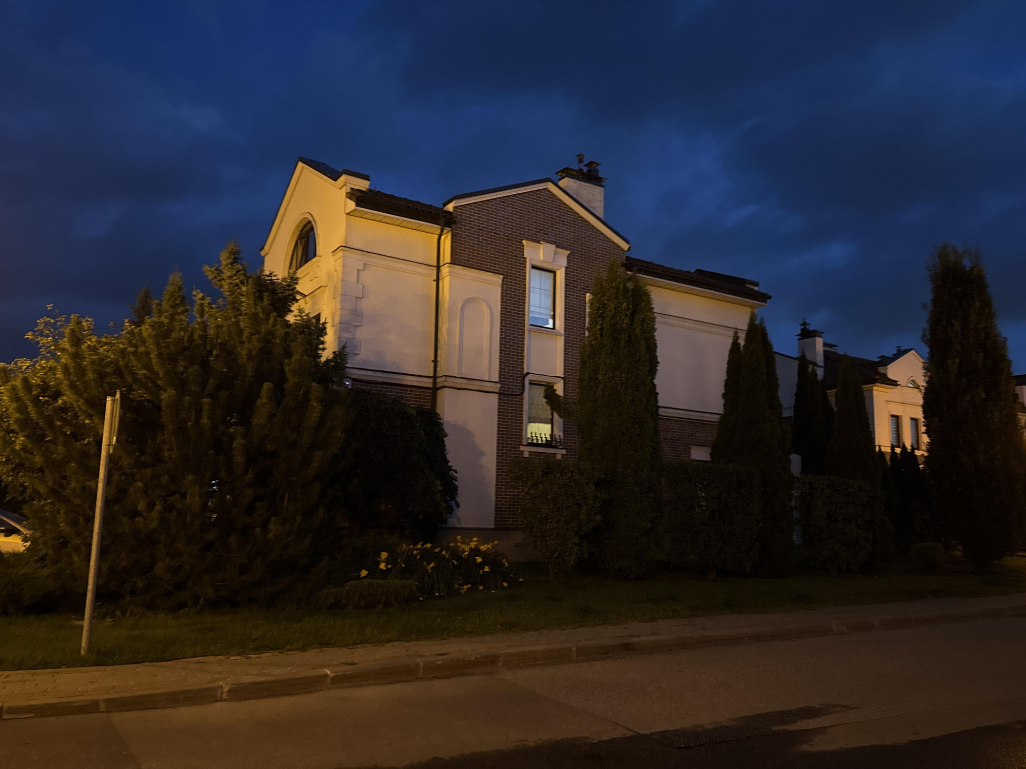 The image depicts a residential street at dusk in Lopatino, Russia. A two-story house with a pale yellow facade and brick accents is prominently featured on the right side of the frame. The house has multiple windows, some of which are illuminated from within, and a prominent chimney. A row of tall, dark green evergreen trees and manicured hedges lines the front of the property, partially obscuring the lower portion of the house. In the foreground, a paved road and a sidewalk are visible, with a metal signpost on the left side of the image. The sky is a deep blue with scattered clouds, suggesting the transition from daylight to night. The lighting in the image is artificial, casting a warm glow on the house and the road, while the surrounding vegetation remains in shadow. No people or animals are present in the scene.