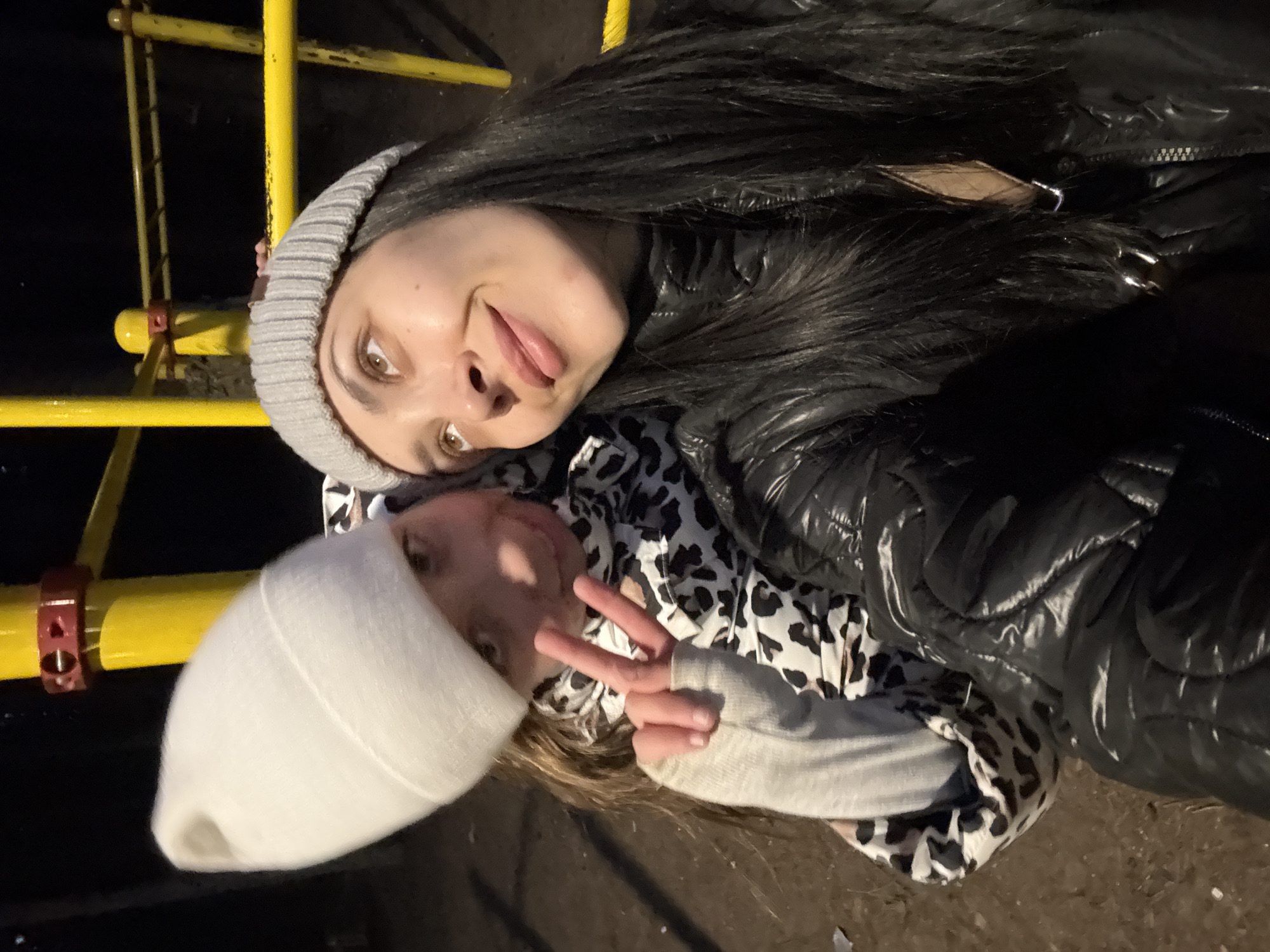 The image features two individuals, likely an adult female and a child, posing playfully for a photograph in what appears to be a dimly lit outdoor setting, possibly a playground in Votkinsk, Russia. The shot is taken from a slightly low angle, with both subjects looking towards the camera.
The adult, positioned above the child, has long dark hair and wears a grey ribbed beanie and a shiny black puffer jacket. She exhibits a playful expression with her tongue slightly out, looking directly at the viewer with brown eyes. Beneath her, the child wears a white beanie and a black and white leopard print top or jacket. The child's hand is raised, making a "peace" sign, and they also appear to be smiling or have a soft, content expression. Both are dressed in warm attire, suggesting cool or cold weather.
The background is mostly dark, indicating it is nighttime. Prominently featured are bright yellow metal bars, resembling a playground structure such as monkey bars or a climbing frame, partially framing the subjects. A small red metal clamp is visible attached to one of these yellow bars. The ground visible below the individuals is dark and uneven. There is no discernible text visible in the image. The overall impression is one of affectionate interaction and joyful, candid moments shared between the two individuals.