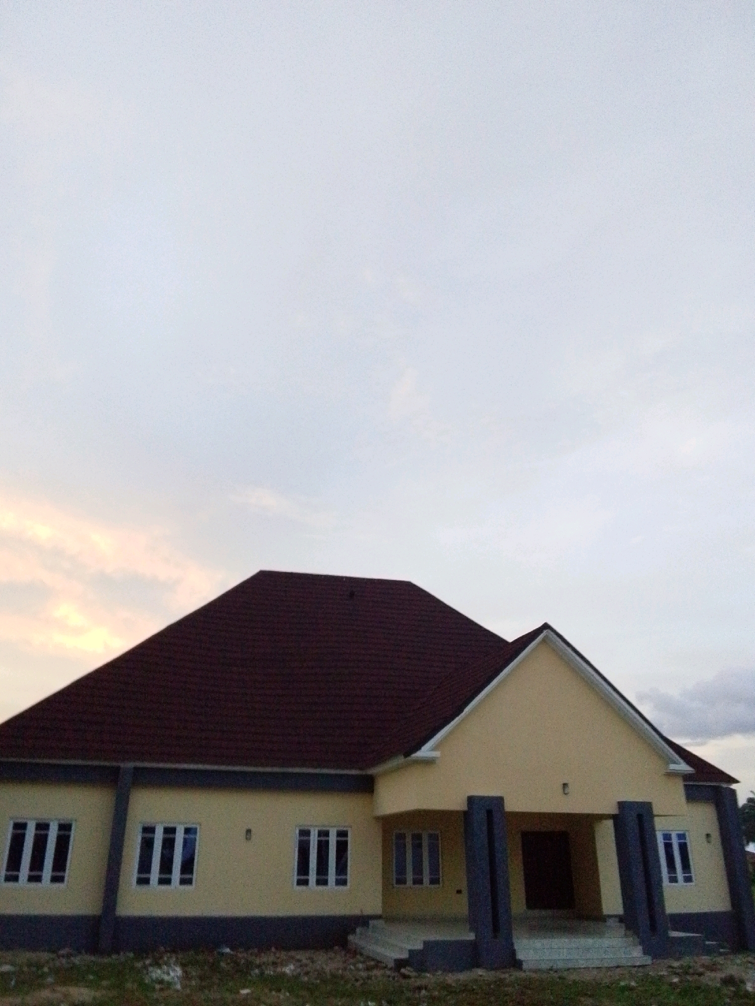 A yellow, single-story house with a dark red roof is pictured under an overcast sky. The house has a modern design with a prominent gabled entrance supported by two dark grey pillars. There are several windows visible, all rectangular with multiple panes. The ground in front of the house appears to be unpaved and covered with sparse vegetation. The image captures the exterior of the residence, with no people or vehicles present. The location is identified as Serti, Nigeria.