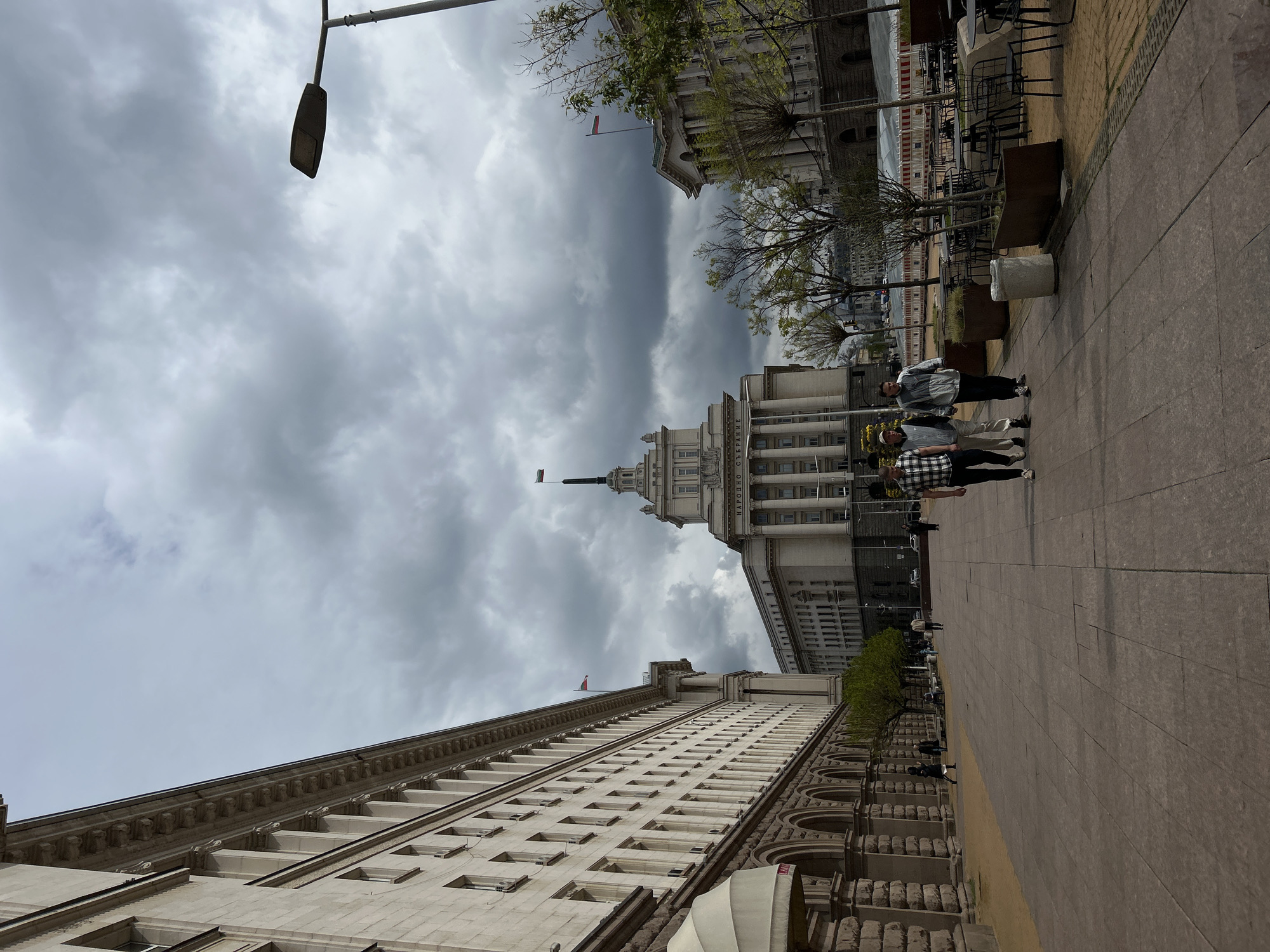 An outdoor, eye-level street view captures a grand urban scene in Sofia, Bulgaria, under a heavily overcast, cloudy sky. The perspective looks down a wide, paved pedestrian area flanked by imposing, monumental buildings characteristic of Stalinist architecture or Neoclassical styles.
On the left, a multi-story building stretches along the frame, featuring numerous rectangular windows and ornate classical detailing, including cornices and horizontal stone bands. On the right, a similar grand building is partially visible, with trees and outdoor furniture in front of it. Further down this side, the distinct building of the Presidency/Council of Ministers stands prominent, characterized by its tall central spire topped with a Bulgarian flag, and a facade adorned with large columns. Another flag, likely Bulgarian, is visible atop the building on the far left.
A group of three casually dressed people walks along the paved sidewalk on the right side of the frame. In the mid-ground on the right, there are outdoor cafe tables and chairs, and some construction barriers, suggesting a public square or renovated area. A streetlight extends into the upper left of the frame.
The overall impression is of a city center on a cool, possibly damp, cloudy day. Visible Cyrillic text on the building with columns on the right reads "НАРОДНО СЪБРАНИЕ" (Narodno Sabranie), confirming it as the National Assembly (Parliament) building in Sofia.