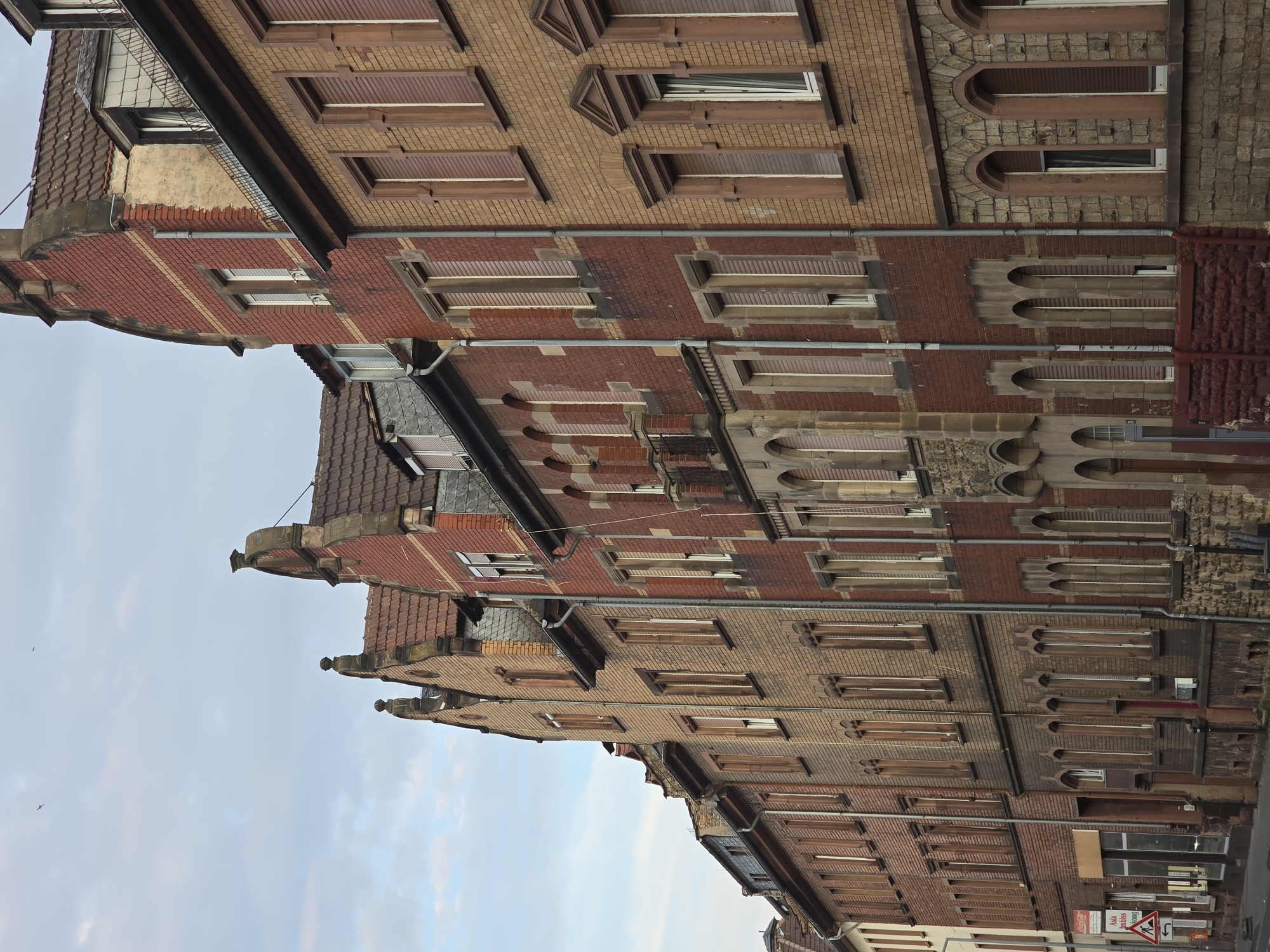 The image shows a row of brick buildings in Worms, Germany. The buildings are several stories high and have various architectural styles, including some with arched windows and gabled roofs. The buildings are made of red and tan brick, and many of the windows are covered with shutters. There is a street in front of the buildings, and a few traffic signs are visible. The sky is cloudy. The scene appears to be a residential or commercial street in a historic part of the city.