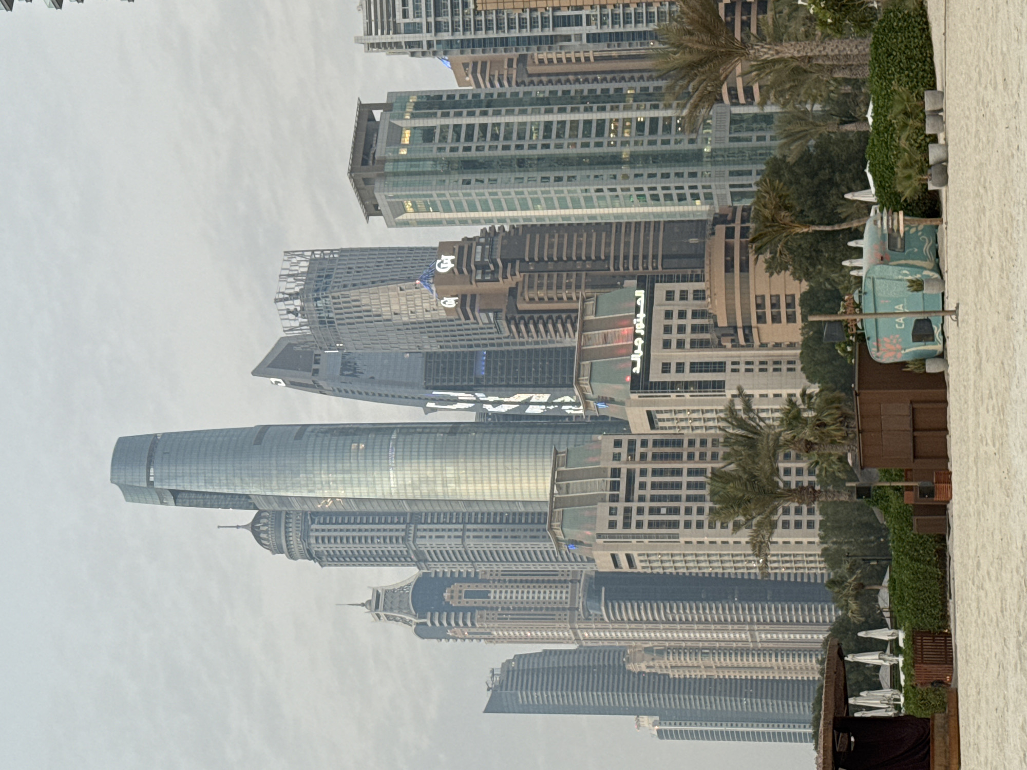 This image captures a view of the Dubai Marina skyline from a sandy beach. The foreground shows a sandy beach with several beach umbrellas and a wooden structure. In the midground, there are palm trees, lush green bushes, and a turquoise-colored food truck or cart with the word "CAVA" printed on it. Behind the beach amenities, a cluster of modern skyscrapers dominates the background, showcasing a mix of architectural styles. The sky is overcast, creating a soft, diffused light. The skyscrapers are of varying heights and designs, featuring glass facades and geometric shapes. Some buildings appear to be residential or hotel towers, while others are clearly office buildings. The overall scene depicts a developed urban coastal area in Dubai, United Arab Emirates.