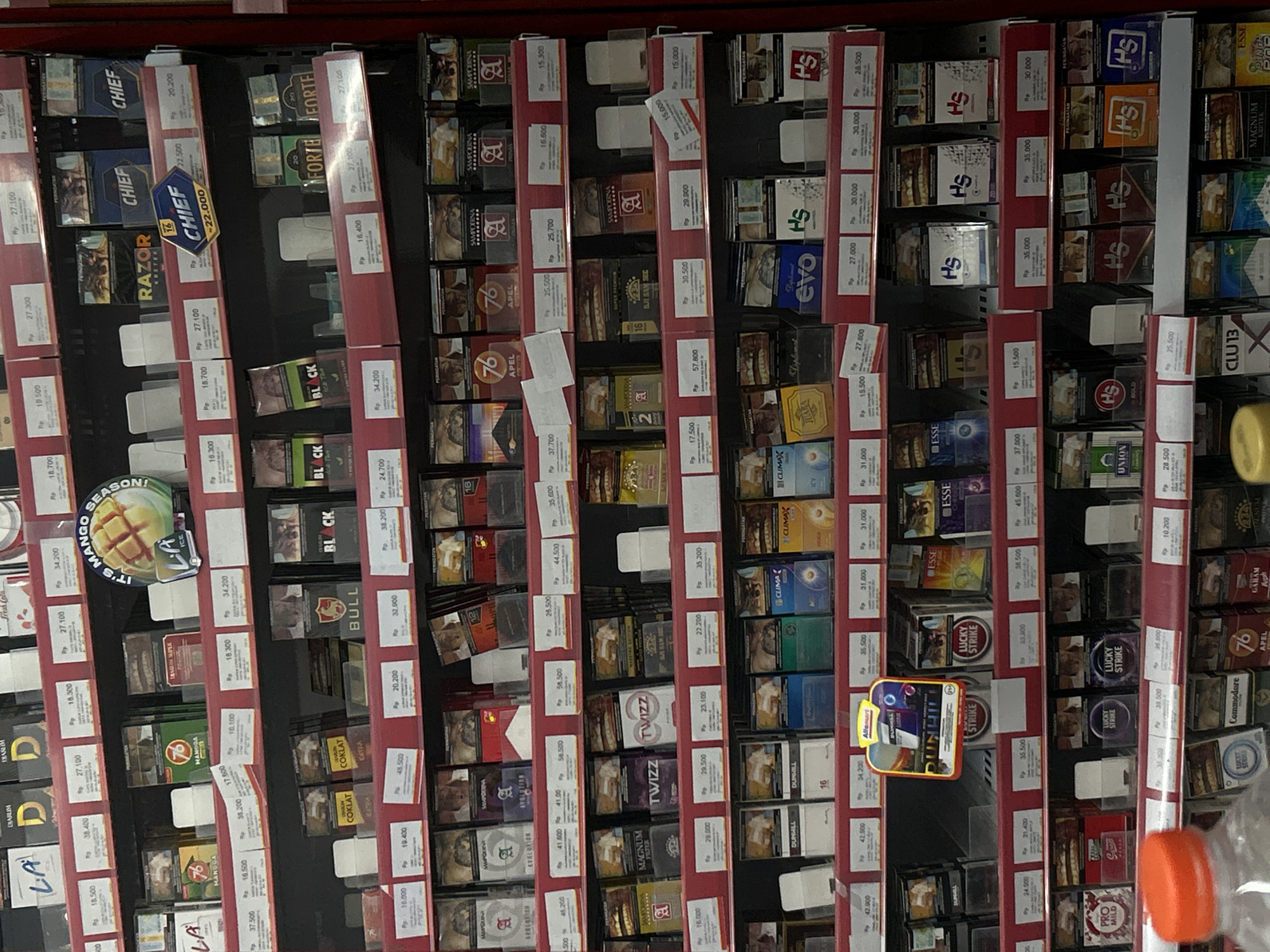 This image captures a vertical display of numerous cigarette packs arranged neatly on shelves within what appears to be a store or minimarket in Payadua, Indonesia. No people are visible.
The shelves are packed with various brands of cigarettes, including local and international labels. Prominently visible brands include CHIEF, RAZOR KRETEK, BLACK, BULL, COKLAT EXTRA, SAMPOERNA A, SAMPOERNA AVOLUTION, 76 APEL, DIARUM LA, TWIZZ, MAGNUM FILTER, BJI SAM SUE, Diplomat, evo, HS, ESSE, LUCKY STRIKE, DUNHILL, CLIMAX, PRO MILD, UNION BOLD, CLU 13, GARAM APH, and Commodore. Many packs feature "PERINGATAN" (warning) text, indicating health warnings commonly found on tobacco products.
Each cigarette pack is accompanied by a price tag displaying its cost in Indonesian Rupiah (Rp). Prices range from Rp 10.200 to Rp 58.500, with many prices in the Rp 15.000 to Rp 35.000 range. A notable marketing display reads "IT'S MANGO SEASON!" with a "LA ICE" logo, possibly indicating a flavored variant. Another small advertising element shows "BUNGHIL POP." A yellow plastic bottle is partially visible on the right side of the frame. The overall scene depicts a well-stocked retail display of tobacco products.