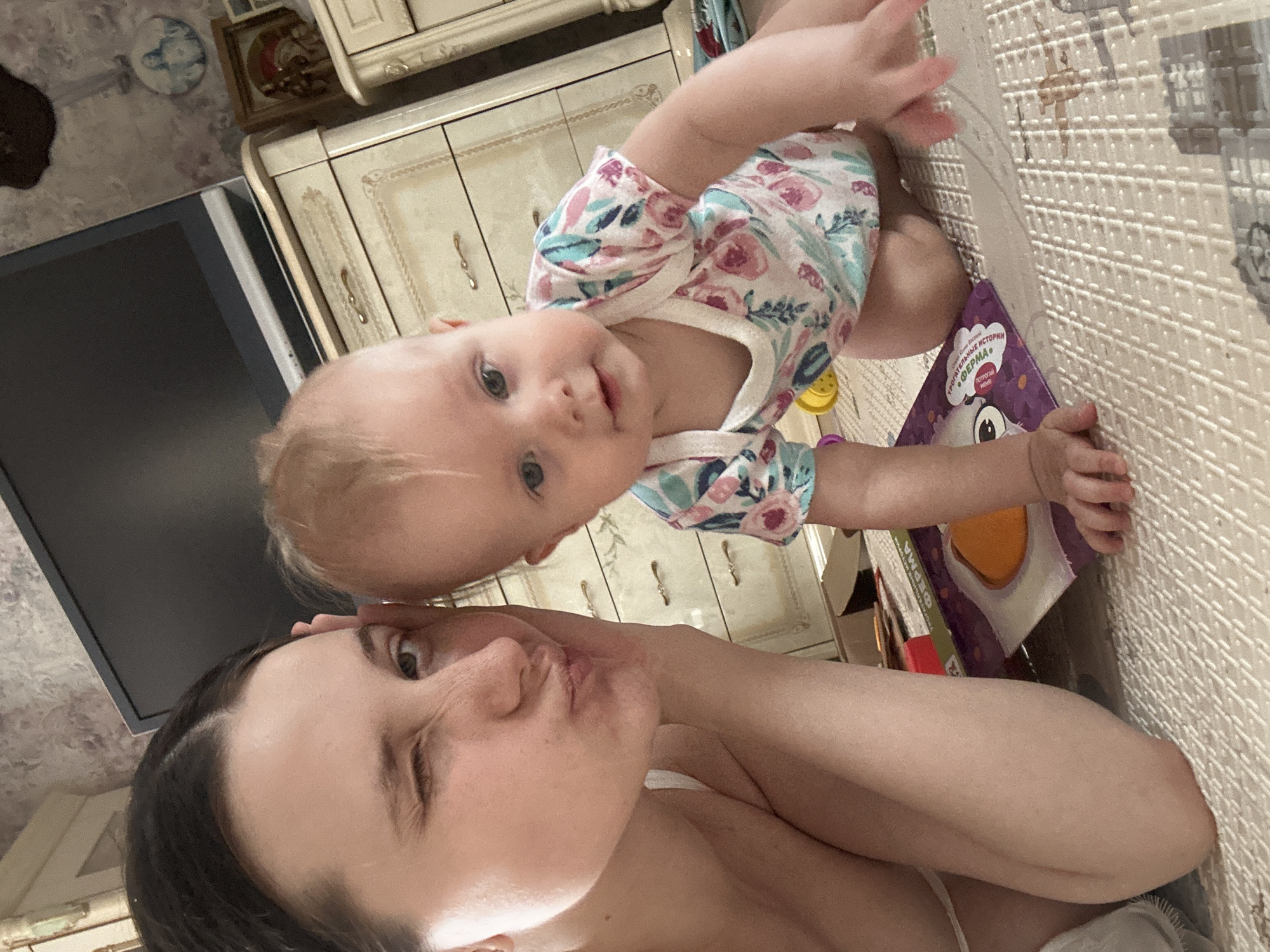 The image depicts a selfie taken indoors, likely in Pokrov, Russia, featuring a woman and a baby. The woman is fair-skinned with dark hair pulled back. The baby, also fair-skinned, wears a light-colored outfit with a floral pattern. The baby is positioned above and slightly behind the woman, looking towards the camera. A book with Russian text and a cartoon-like illustration is near the baby's hand. The background includes a light-colored cabinet with decorative details, a dark-colored television, and a patterned floor mat.
