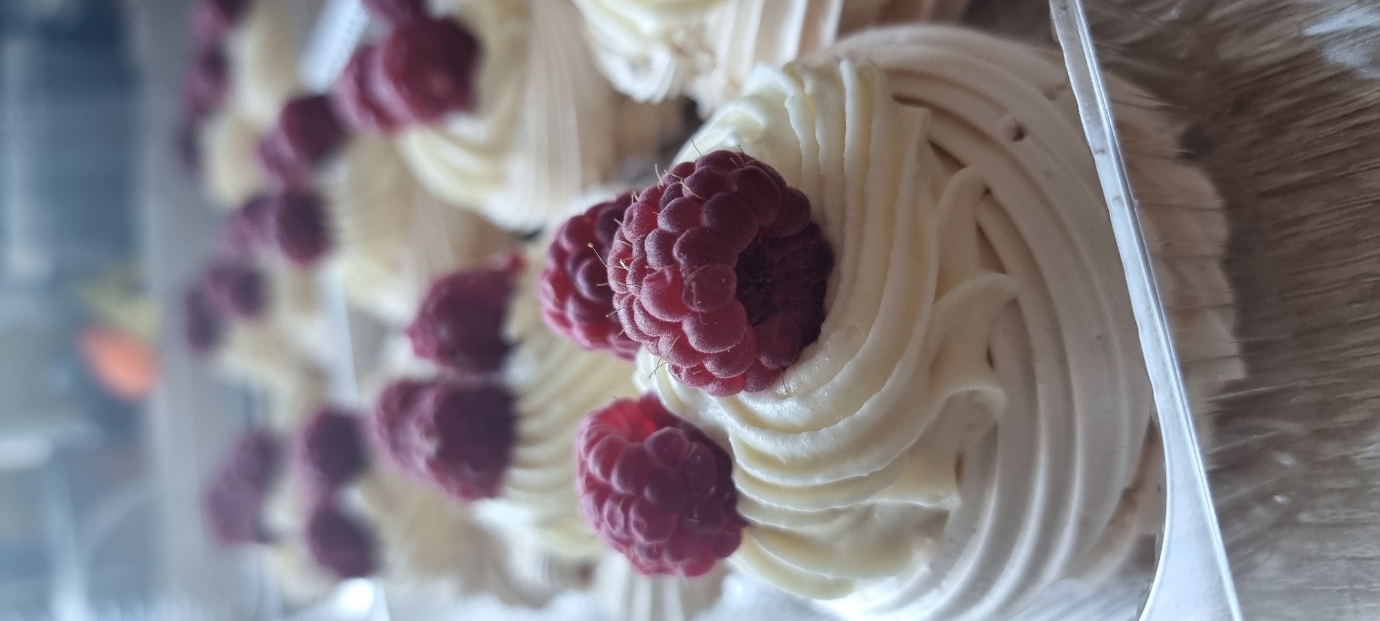 The image shows a close-up, vertical shot of several mini pavlovas with whipped cream and raspberries. The pavlovas are arranged in a shallow, rectangular aluminum foil container. The pavlova in the foreground is in sharp focus, showcasing the intricate swirls of whipped cream and the textured surface of a ripe raspberry. Behind it, other pavlovas are visible, some with raspberries and some appearing to be plain. The background is out of focus, showing a muted, indistinct scene that could be an indoor setting. There are no people or specific landmarks of Aktobe, Kazakhstan, visible in the image.