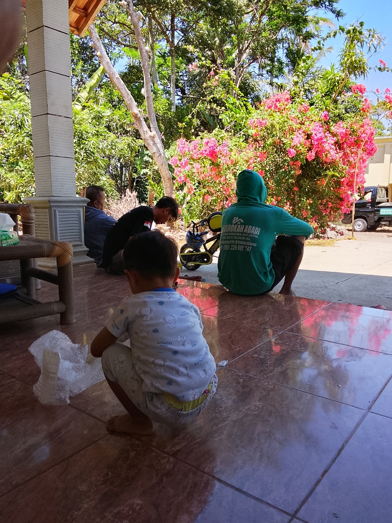 In Kradenan, Indonesia, a group of four individuals are gathered on a tiled porch in front of a building with a wooden overhang. A young child in a white shirt with elephant patterns is squatting in the foreground, facing away from the camera. To their left, a roll of bubble wrap is placed on the floor. Behind the child, three adult males are seated. One man, wearing a dark shirt, is looking down at something on the ground. Another man, wearing a blue robe, is seated further back. A third man, wearing a green hooded sweatshirt with text that reads "KAROKAH ABADI RUMAH BANGUNAN ALAT LISTRIK," is also seated, facing away from the camera. A bicycle and a black pickup truck are visible in the background. The scene is illuminated by bright sunlight, casting shadows on the porch and revealing lush greenery and pink flowers surrounding the area.