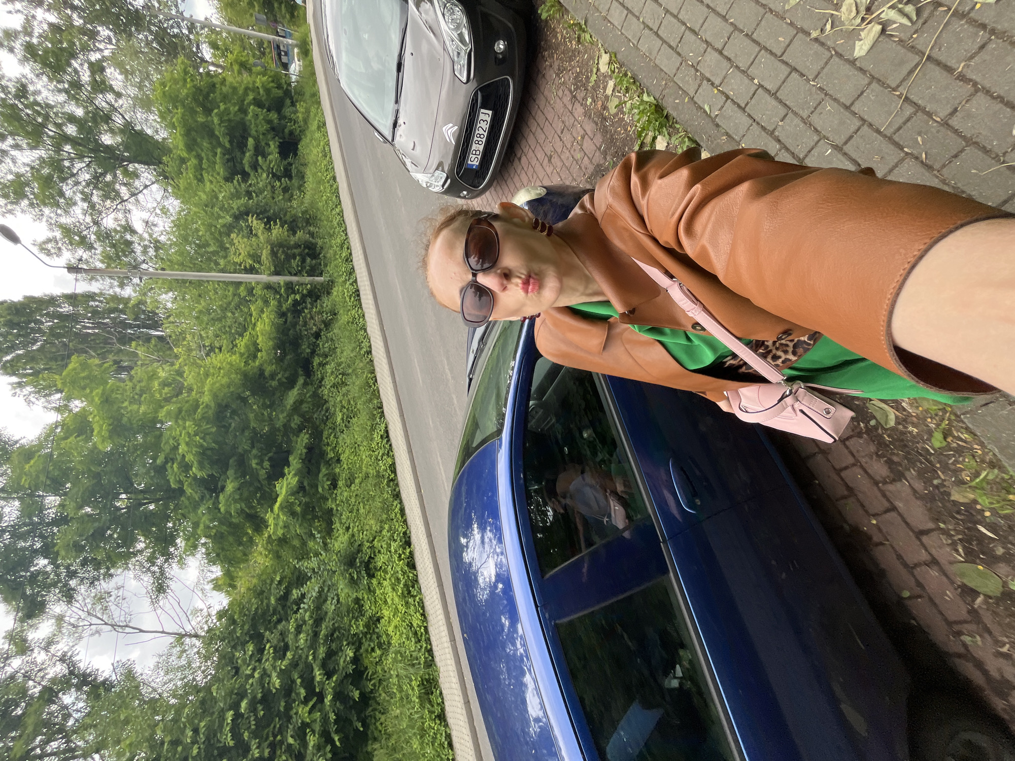In a medium shot, a woman stands on a sidewalk next to a parked blue car, with another brown car parked further down the street. She is wearing a brown leather jacket, a green shirt, and sunglasses, and she is puckering her lips towards the camera. A pink purse hangs over her shoulder. The scene appears to be in an urban or suburban setting, with trees lining the street and a cloudy sky overhead. The license plate of the brown car reads "SB 8823 J".