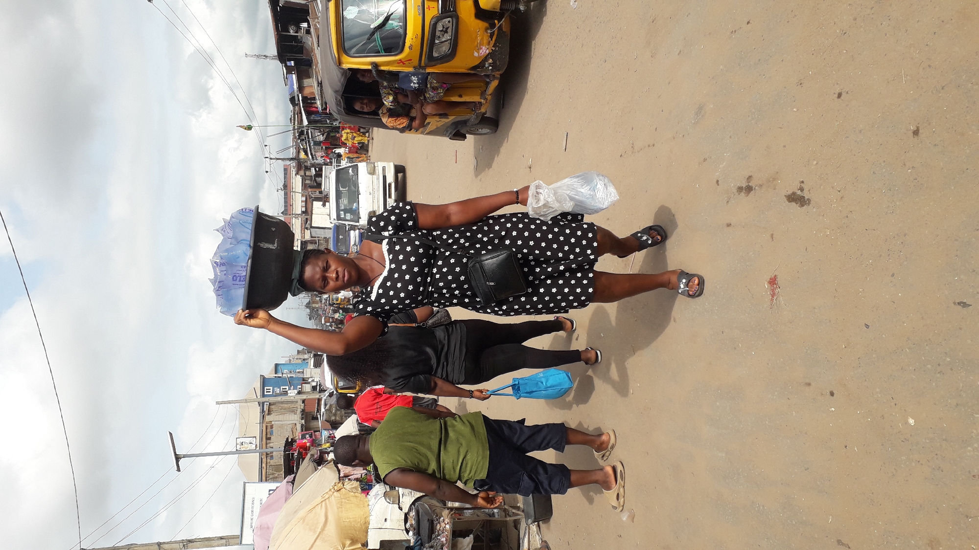 The image depicts a street scene in Onitsha, Nigeria, likely a market area. In the foreground, a woman is walking towards the viewer, balancing a large, dark basin filled with blue-wrapped items on her head. She is wearing a black and white floral print dress and black sandals, and she carries a plastic bag in her right hand. Behind her, other people are visible. To her left, a man in a green sleeveless shirt and dark shorts walks away from the camera, and to her left and further back, a woman in black walks away carrying a blue bag. In the background, various vehicles are present, including a white van and a yellow three-wheeled auto-rickshaw with passengers. Stalls and buildings line the street, suggesting a commercial environment. The ground is paved with a mixture of dirt and asphalt, and the sky is overcast with clouds.