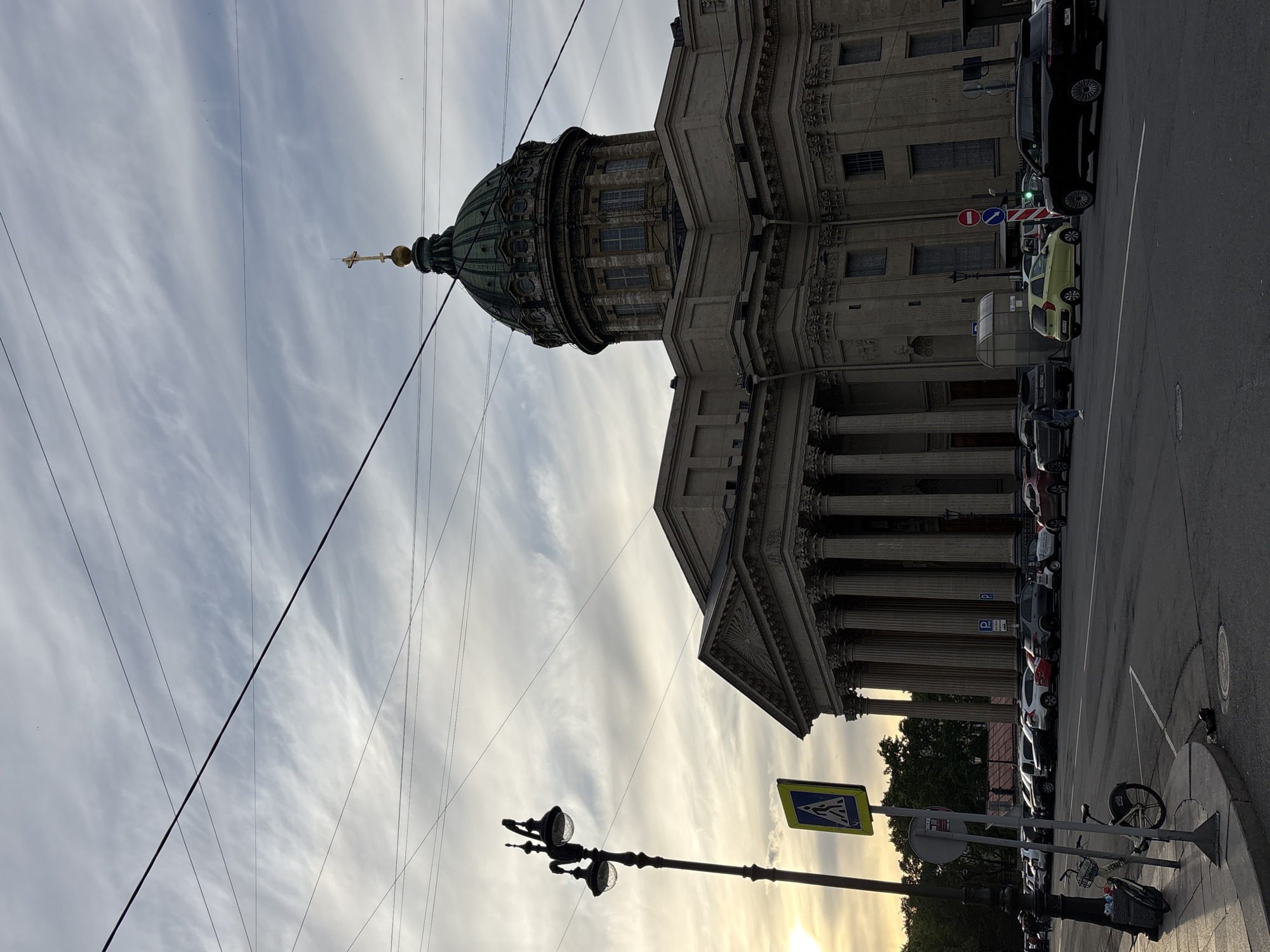 The image, captured in Saint Petersburg, Russia, depicts the grand façade and distinctive dome of a large, classical-style cathedral, likely the Kazan Cathedral. The building features numerous fluted columns and elaborate architectural details, characteristic of Neoclassical design. A golden cross, indicative of a religious structure, surmounts the green-patinated copper dome, which is a prominent feature against the sky.
The scene is set on an urban street during what appears to be either early morning or late afternoon, as suggested by the warm, low sun visible near the horizon and the soft, diffused light illuminating the streaky, cirrus clouds. A network of dark power lines stretches across the cloudy sky.
Along the street running parallel to the cathedral, a row of cars is parked. A vintage-style black street lamp with two clear globes stands in the foreground. Visible road signage includes a blue pedestrian crossing sign with a white walking figure, a red circular 'No Entry' sign with a white horizontal bar, a blue circular 'No Stopping' sign with a red diagonal line, and a blue square parking sign marked with a white 'P'. A bicycle is also parked on the sidewalk. No explicit activity involving people is visible, but the presence of vehicles and street furniture points to an active urban environment.