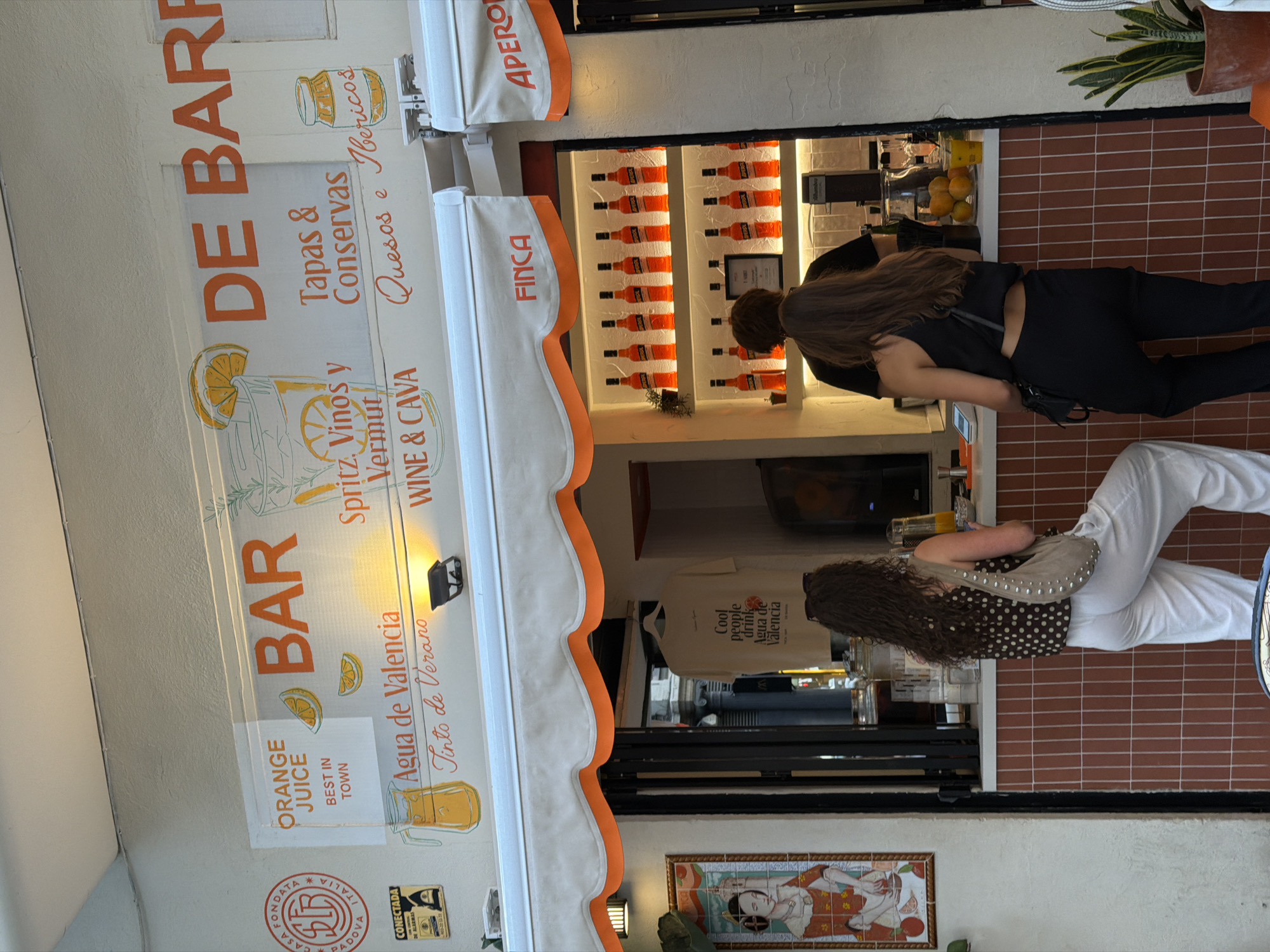 This media file captures an outdoor scene at what appears to be a bar or cafe. The primary subjects are two individuals, both women, interacting with the establishment. One woman, wearing black attire and carrying a bag, is standing at a counter with bottles of orange liquid displayed behind her. The other woman, in white pants and a patterned top, is also at the counter, facing away from the camera.
The setting is a storefront with a striped awning overhead, displaying the words "DE BARR" and "BAR" in large orange lettering. Various menu items and offerings are advertised on the awning, including "Tapas & Conservas," "Quesos e Ibericos," "Spritz. Vinos y Vermut," and "WINE & CAVA." There are also indications of drinks like "ORANGE JUICE" and "Agua de Valencia." The overall ambiance suggests a vibrant and possibly daytime setting, with warm lighting and the presence of beverages.
Visible text includes numerous Spanish and Italian words related to food and drink. Notably, "FINCA" and "APERO" are also displayed on the awnings. A framed artwork with what looks like traditional Japanese or Asian figures is visible near the bottom of the frame. The presence of multiple beverage bottles and the counter suggest a commercial establishment focused on serving drinks. The image is taken from a slightly low angle, looking upwards towards the awnings and the interior of the bar. The overall mood seems casual and inviting, typical of a relaxed establishment.