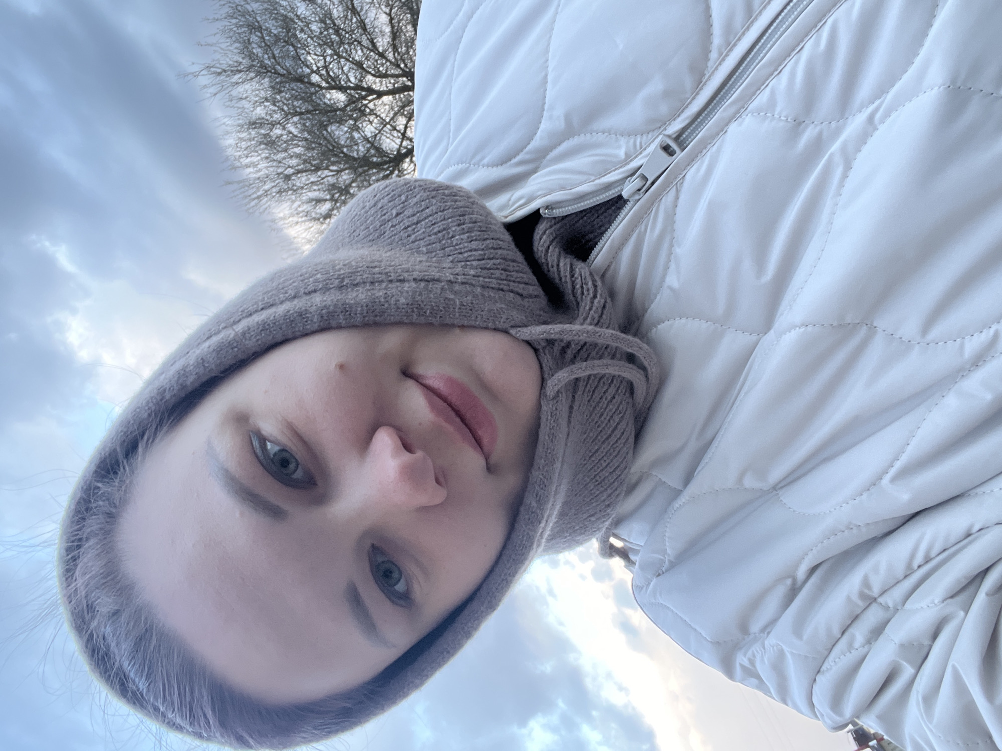 The image depicts a woman with fair skin, wearing a light beige puffer jacket and a knitted hood, looking into the camera. The background shows a sky with cloudy patterns and bare tree branches. The photo seems to be taken from a low angle, pointing upwards. Given the information provided, the location is Usman, Russia.