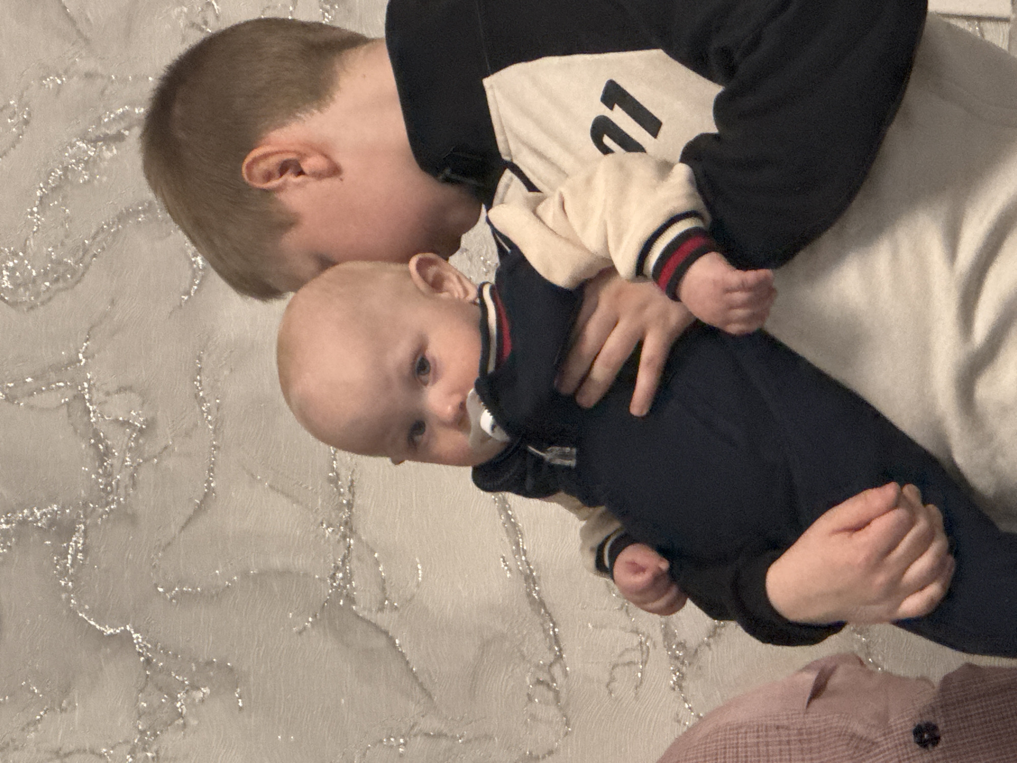 This image captures an intimate indoor moment between two individuals in Chaikovsky, Russia. An older child, likely a boy with short light brown hair, is seen holding an infant. The older child is wearing a dark blue and white sweatshirt featuring a large black number "21" on the white section. He is leaning his head towards the infant, suggesting a tender kiss or affectionate nuzzle.
The infant, with fair skin and light hair, is dressed in a dark blue outfit, possibly a onesie, with prominent striped cuffs in red, white, and blue. A white pacifier is visible in the infant's mouth, and they are looking towards the viewer with a calm expression. The older child's hands are gently supporting the infant.
The background consists of a light-colored wall adorned with a shimmering silver patterned wallpaper. A glimpse of pink and white plaid fabric is visible in the lower right corner, suggesting another piece of clothing or a blanket. The lighting is soft, indicative of an indoor setting, likely a home. The scene conveys a strong sense of affection, warmth, and familial bonding between the two.