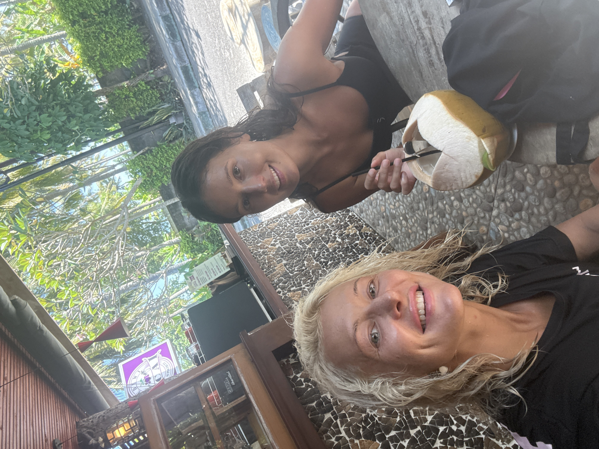 Two women are pictured outdoors at a table. The woman on the left has dark hair and is holding a coconut with a spoon in it. She is wearing a black top. The other woman has blonde hair and is wearing a black shirt. A backpack sits on the table. The table surface has a mosaic tile pattern. Trees and a building are visible in the background. The city and location are unknown.
