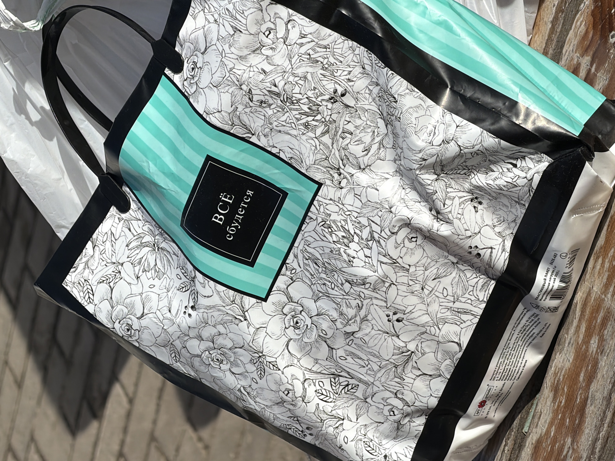 A plastic shopping bag sits on an outdoor surface, possibly a wooden bench or table. The bag has a floral pattern in black and white, with a teal panel featuring white stripes and a black square with the text "ВСЁ сбудется" in white Cyrillic letters. The bag has black handles and trim. Text in Russian and English near the bag's bottom edge indicates it is a plastic bag, with the Importer listed as "ARTICENTER" in Moscow. The background is a slightly blurred view of what appears to be a sidewalk or paved area.