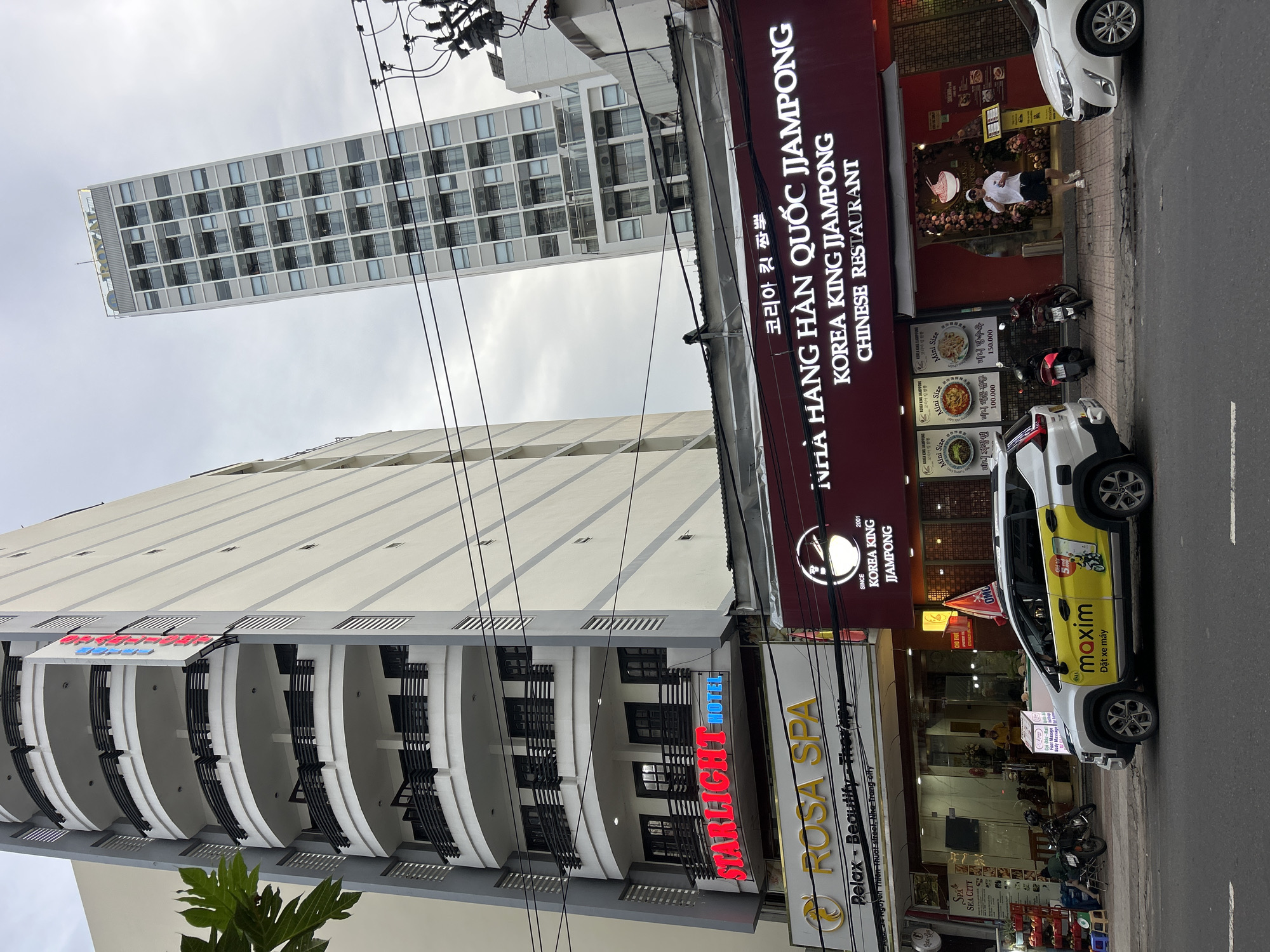 An urban street scene in Nha Trang, Vietnam, features several commercial buildings under an overcast sky. In the foreground on the left, a multi-story building houses the "STARLIGHT HOTEL," identified by a prominent red and blue sign. Below it, a "ROSA SPA" sign in gold lettering advertises "Relax - Beautify - Therapy" and provides the address "34 Nguyen Thien Thuat street, Nha Trang city." Another small sign for "SPA SEA CITY" is also visible.
Adjacent to these establishments, a dark red signboard for a restaurant reads "NHÀ HÀNG HÀN QUỐC JJAMPONG," "KOREA KING JJAMPONG," and "CHINESE RESTAURANT" in Vietnamese, English, and Korean ("코리아 킷 짬뽕"). Smaller signs below showcase "Mini Size" dishes, including "Mini Jajangmyeon," "Mini Seafood Jjamppong," and "Mini Tangsuyuk," with prices of 100,000 and 150,000, likely Vietnamese Dong. A person in a white t-shirt stands outside this restaurant.
On the street, a white SUV branded with "maxim" and "Đặt xe máy" (Order motorbike) in yellow text, along with a "5%" offer, is parked by the curb. Several motorbikes are also parked nearby. In the background, a tall, modern high-rise building with numerous windows extends into the cloudy sky. A dense network of black power lines crisscrosses the upper part of the image. The scene depicts a typical commercial area in an Asian city, with a mix of local businesses and international influences.