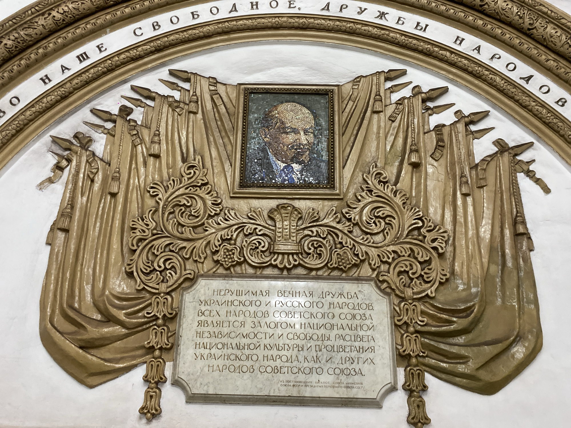 The image captures a decorative wall piece from a Moscow, Russia metro station. At the top, text in Cyrillic script forms an arch. Below, a framed mosaic portrait of Lenin is centered above a stone tablet with Cyrillic text. The portrait and tablet are flanked by sculpted gold-colored drapes. Additional gold-colored ornamentation surrounds the portrait and tablet.