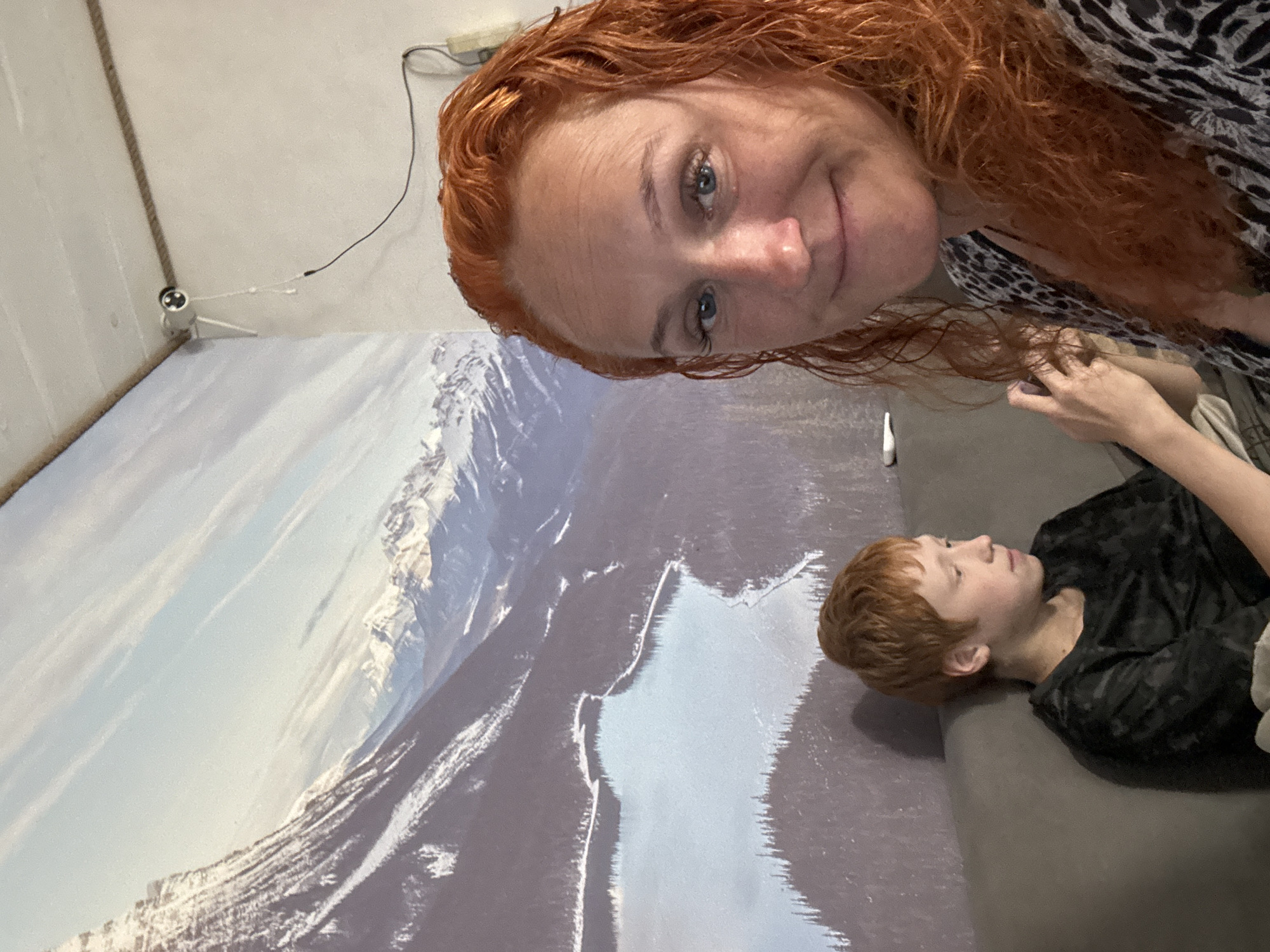 The media file features a woman with long, wavy red hair taking a selfie with a young boy. The woman is positioned in the foreground, leaning in towards the camera with a smile. She is wearing a patterned top. The boy is lying on a sofa in the background, wearing a dark patterned shirt. Behind them is a large mural or backdrop depicting a snowy mountain landscape with a lake or river. On the wall above the mural, a small white security camera is visible, connected to a wire. The scene appears to be indoors, possibly in a home or studio. The lighting is natural, suggesting daytime. The overall mood is casual and intimate, with both subjects looking relaxed. No text is visible in the media file.