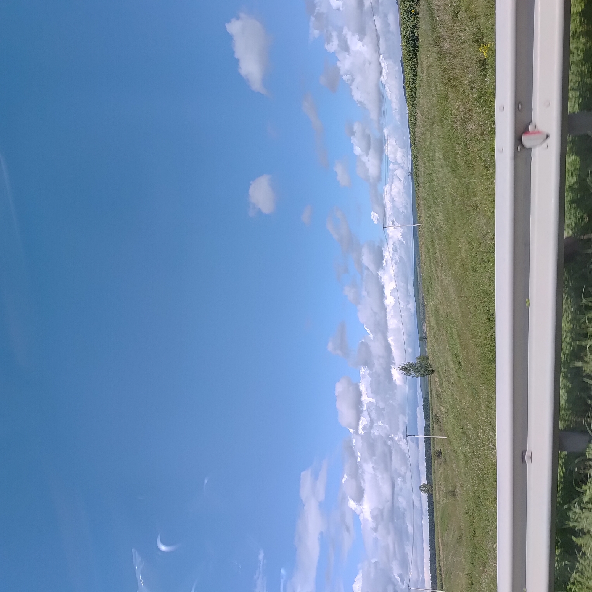 The image depicts a rural landscape under a bright blue sky with scattered white clouds. In the foreground, a silver guardrail runs horizontally across the bottom of the frame. Beyond the guardrail, a green field stretches into the distance, dotted with sparse trees and vegetation. Several utility poles with wires are visible in the mid-ground, traversing the field. In the far background, a line of trees forms a dark green horizon, with rolling hills or mountains beyond. The overall impression is one of a serene, open countryside, possibly taken from a moving vehicle on a road near Staritsino, Russia.