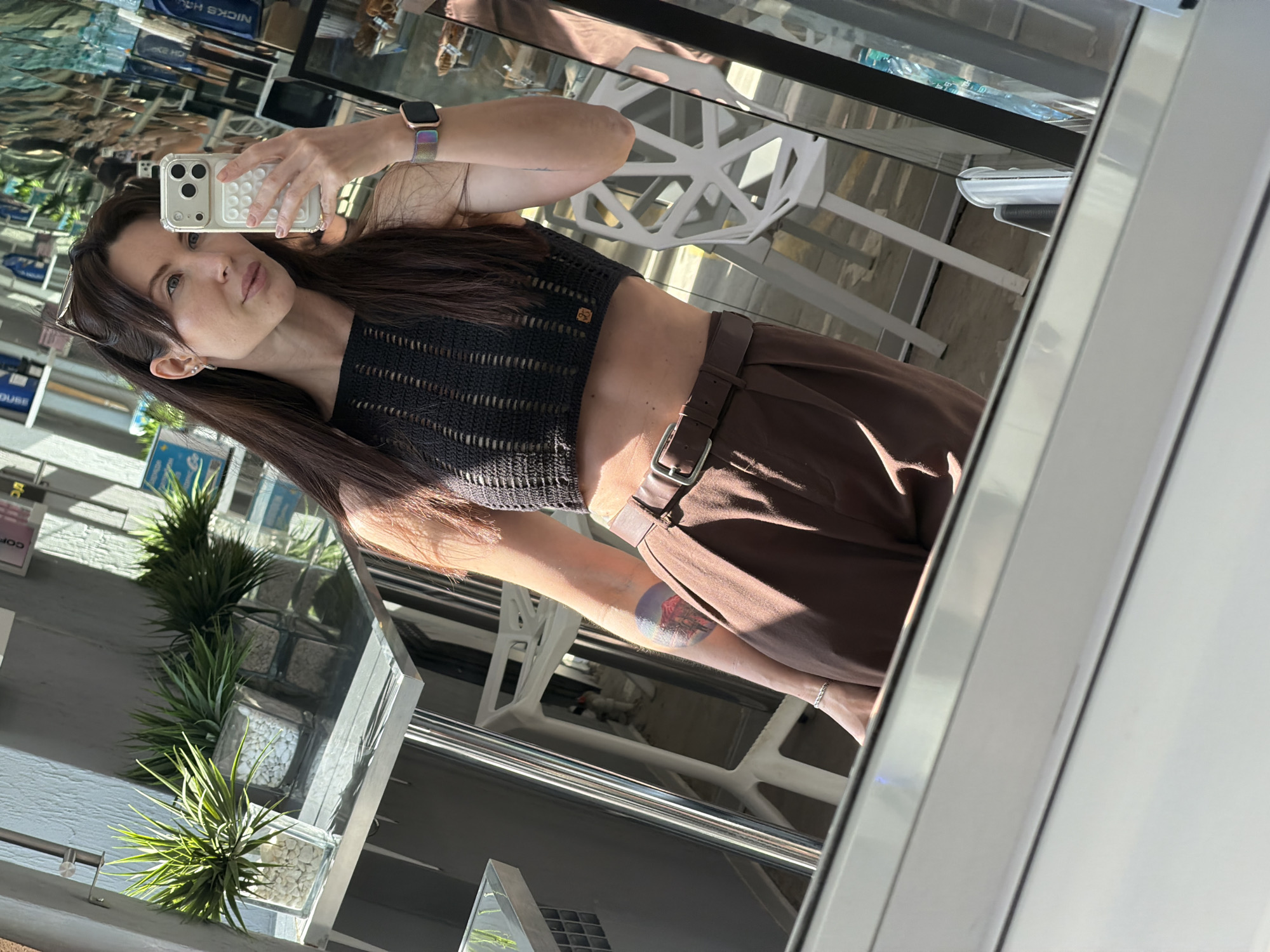 A woman is seen in a mirror reflection, possibly in Perm, Russia given the "Nicks" sign reflected in the mirror. She is holding a white phone and wearing a dark knit top, brown belted pants, and an Apple watch. She has dark hair and fair skin. In the reflection, there's a glimpse of an outdoor setting with plants, tables, and other patrons.