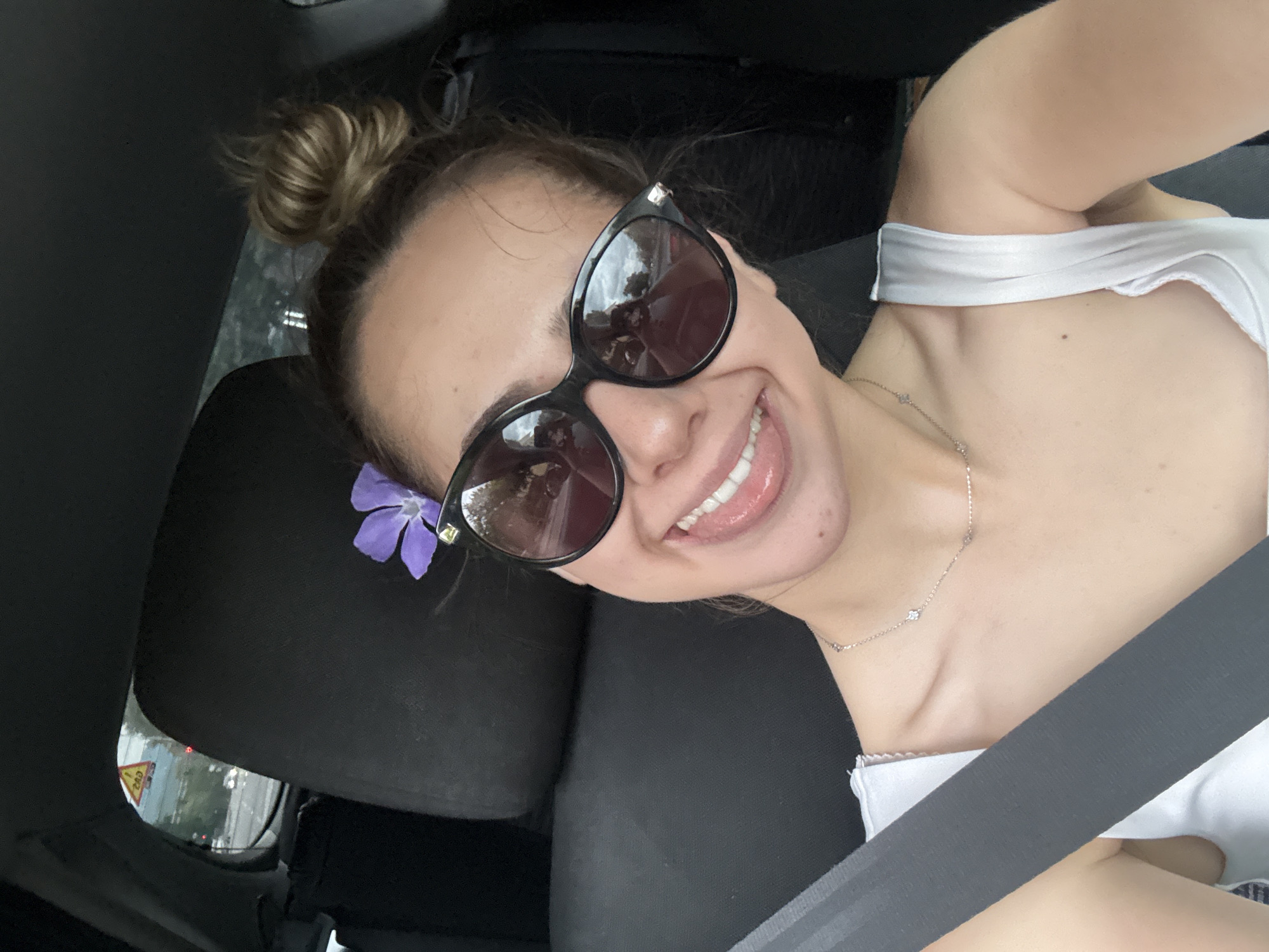 A fair-skinned woman with brown hair styled in a messy bun atop her head is taking a selfie inside a car. She is wearing oversized sunglasses and a white top with a thin silver necklace. A purple flower is tucked behind her left ear, above the sunglasses. She is smiling widely, showing her teeth. A seatbelt is diagonally across her body.
The background indicates she is seated in the driver's seat of a car. Outside the car window, there is a glimpse of an urban environment. A red and white triangular road sign with "200" written on it is visible. Based on the road sign and the general environment, it is likely that the location is in or near Blagoevgrad, Bulgaria. The interior of the car appears to be black.