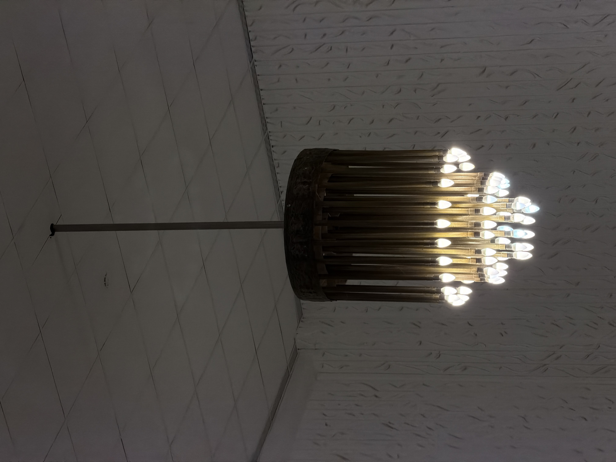 The image shows a chandelier hanging from a ceiling. The chandelier has a circular frame with many cylindrical lights attached. The ceiling appears to have a textured surface. The location is likely within the city of Perm, Russia.