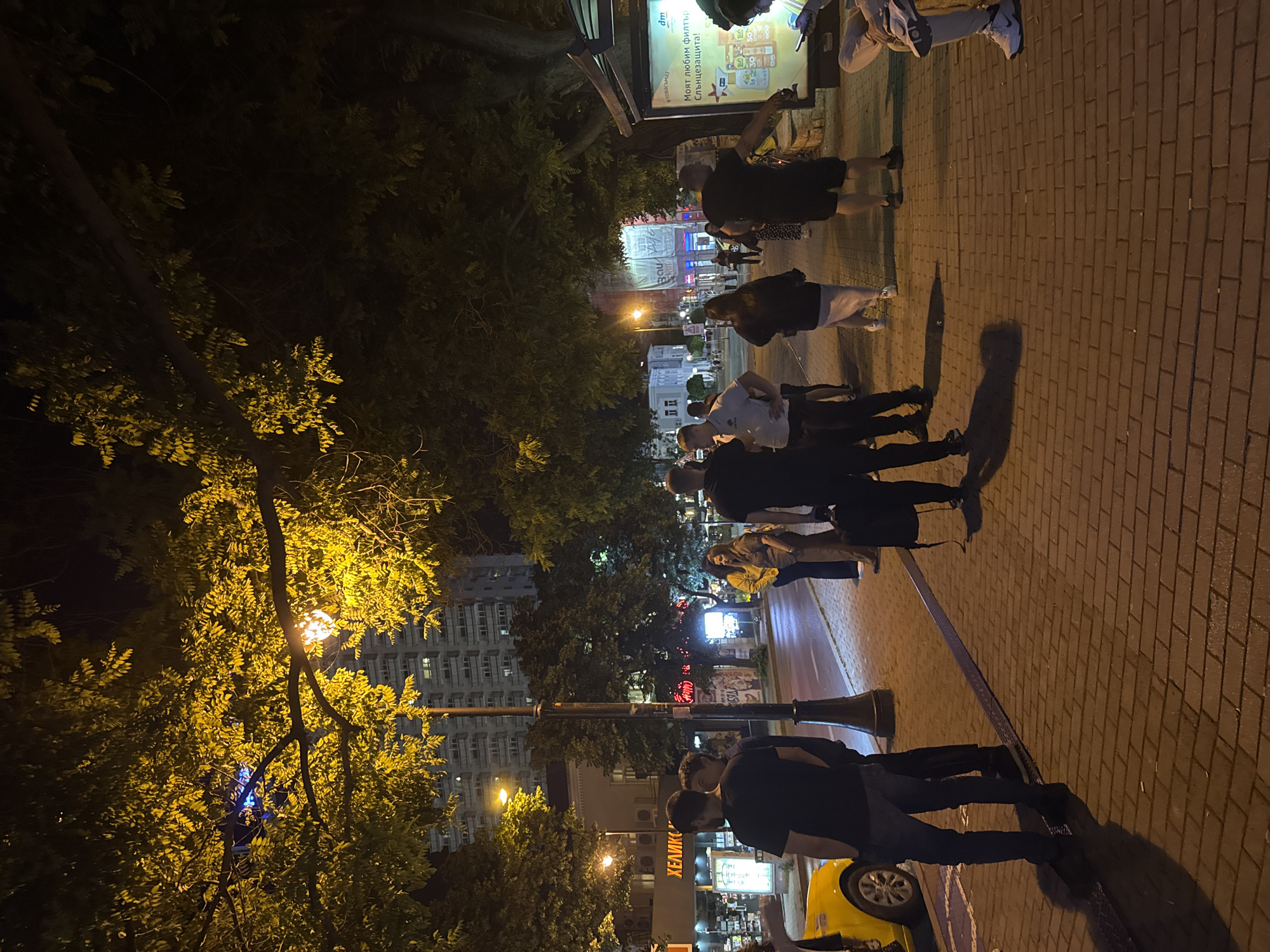 The image depicts a nighttime scene on a street in Varna, Bulgaria, likely near a commercial area given the storefronts and illuminated signs. A yellow taxi is parked on the left side of the street. Several individuals are standing on the brick-paved sidewalk, some in small groups, others individually. The lighting is a mix of streetlights and the warm glow from the trees overhead, creating a somewhat atmospheric, nocturnal urban environment. In the background, a tall residential building with numerous illuminated windows is visible, suggesting the presence of apartments or hotels in the vicinity. A bus stop shelter with an advertisement is present on the right side of the sidewalk, indicating public transport services. The overall impression is of a lively urban setting at night with people engaged in casual social interactions or awaiting transportation.