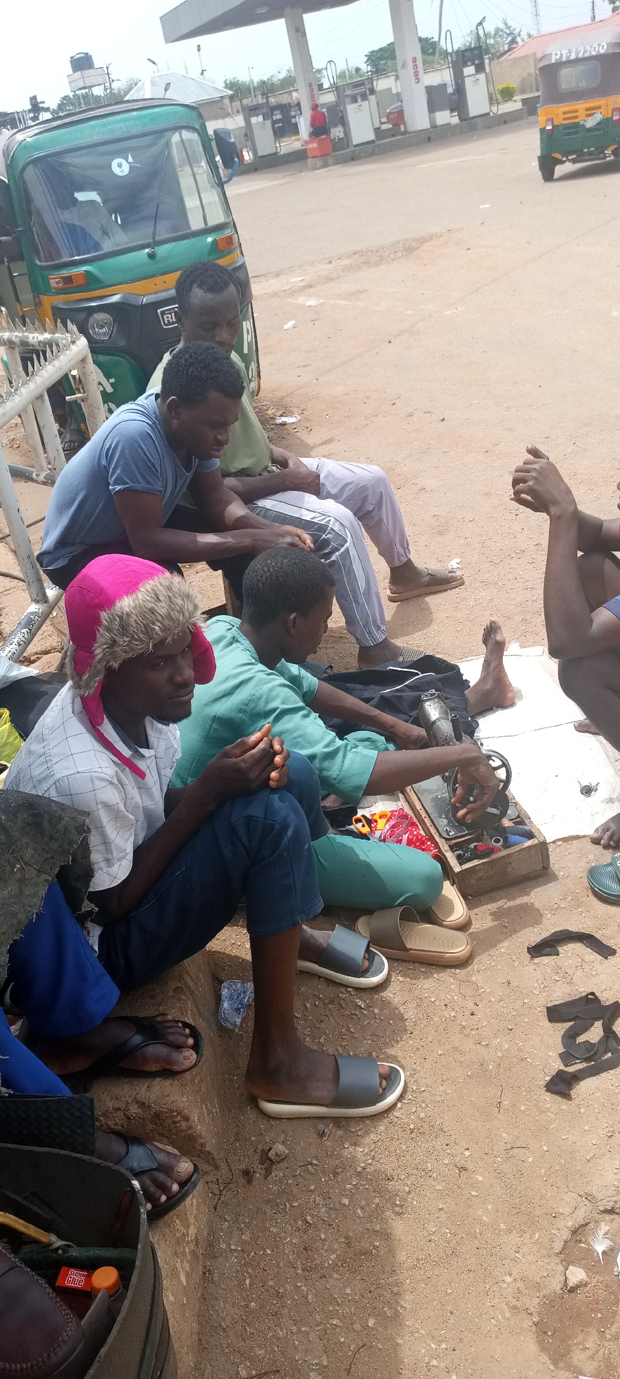 Five men are seated outdoors on the side of a road, with a small, hand-cranked sewing machine positioned on a white cloth on the ground in front of them. Several pieces of black material, likely scraps of leather or fabric, are also on the ground. Some of the men are wearing sandals. An open wooden box contains sewing supplies, and spools of thread. A pink furry hat is worn by one of the men. The road is unpaved. In the background, a green and yellow tricycle taxi is parked next to a white fence. In the middle distance is a gas station. The setting appears to be Gura Top, Nigeria.